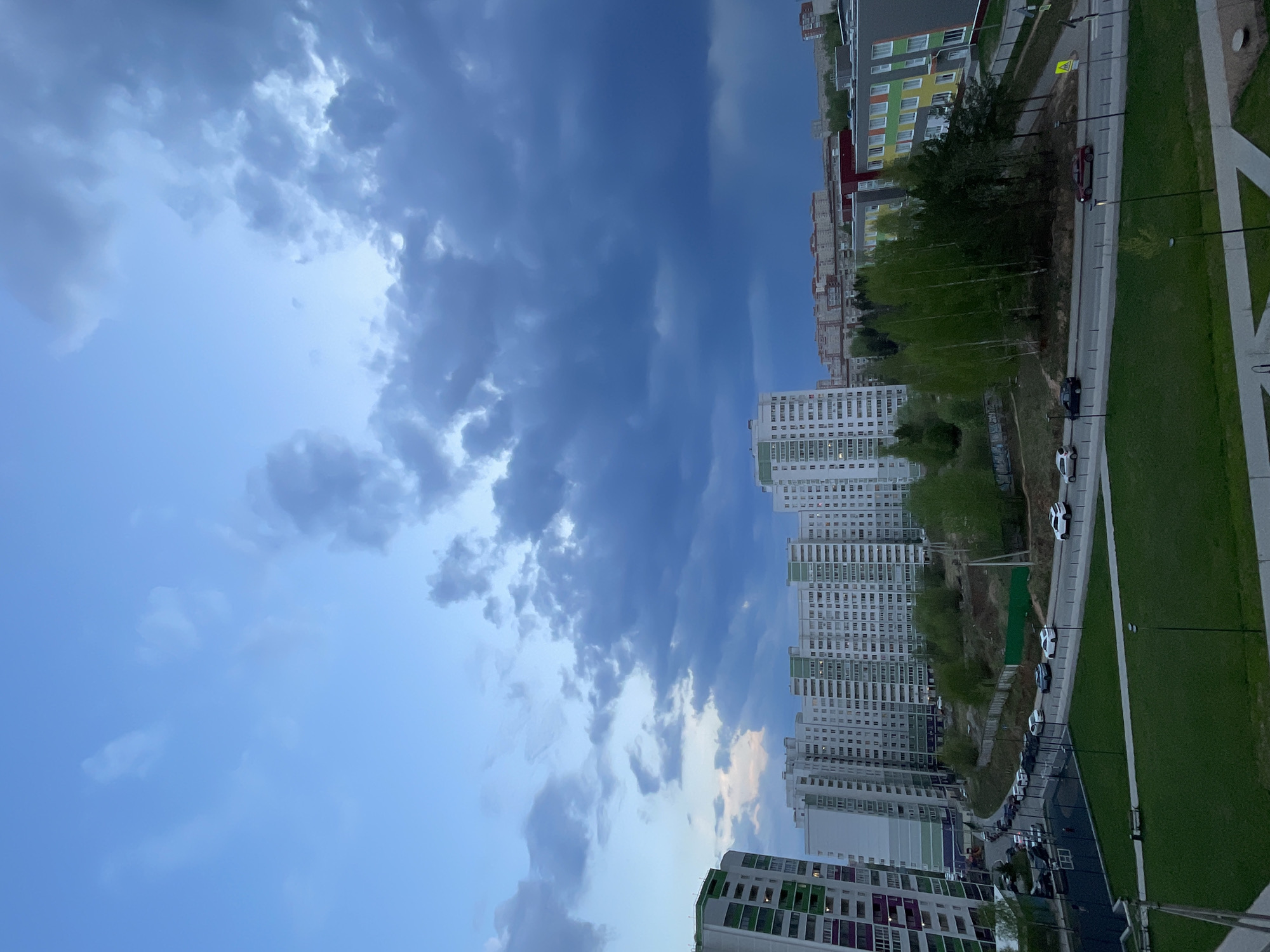 The image depicts a partially cloudy sky over a cityscape, likely taken from a high vantage point. In the foreground, rows of multi-story apartment buildings are visible, painted in various colors. A road runs parallel to the buildings, with parked and moving cars. The sky transitions from lighter blue with scattered clouds on the left to darker, stormier clouds on the right. The cityscape includes green spaces, possibly parks or lawns. The location may be Izhevsk, Russia, but this cannot be confirmed with certainty based on the image alone.