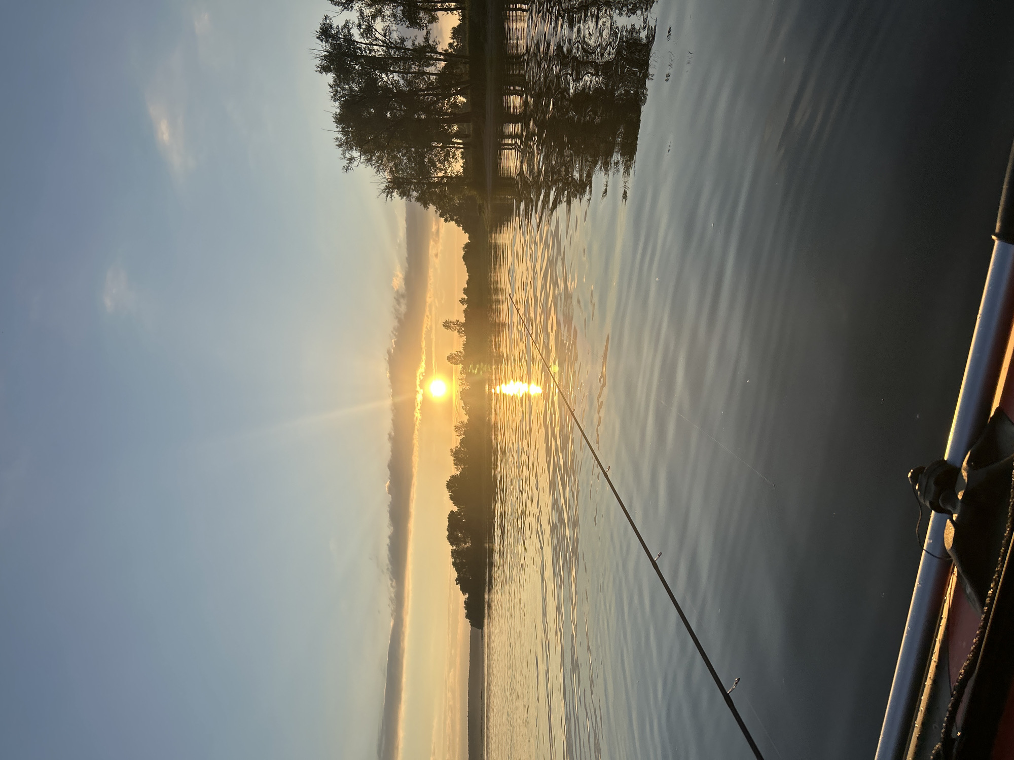This is a photograph of a sunset over a body of water, likely taken from a boat or kayak. The sun, a bright orb, is low on the horizon, casting a warm golden glow across the scene. Rays of sunlight emanate from the sun, illuminating the sky and reflecting off the rippling surface of the water. In the distance, a line of trees forms a silhouette against the illuminated sky, creating a natural barrier between the water and the land. A fishing rod extends from the foreground, its line disappearing into the water, suggesting an angler is present, though no person is visible in the frame. The overall impression is one of tranquility and the beauty of nature at dusk. The location is identified as Ol'khovskoye, Russia, though no specific landmarks confirm this.