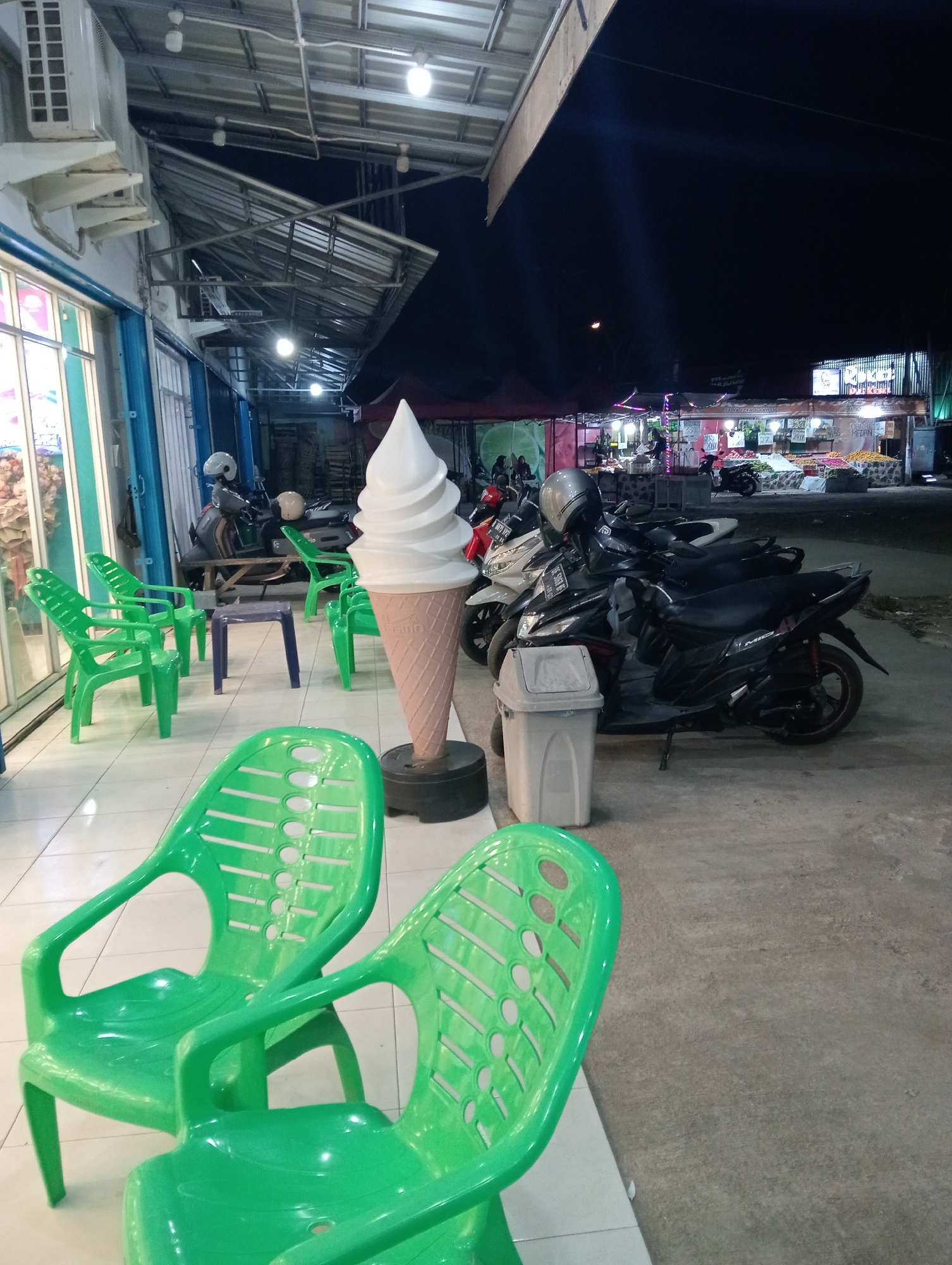 A nighttime street scene in Pringsewu, Indonesia, features an outdoor seating area with several bright green plastic chairs and a tall, cone-shaped ice cream statue. In the background, a row of shops and market stalls are illuminated, displaying various goods. Several motorcycles are parked along the street. The overall atmosphere is that of a bustling night market or roadside eatery.
