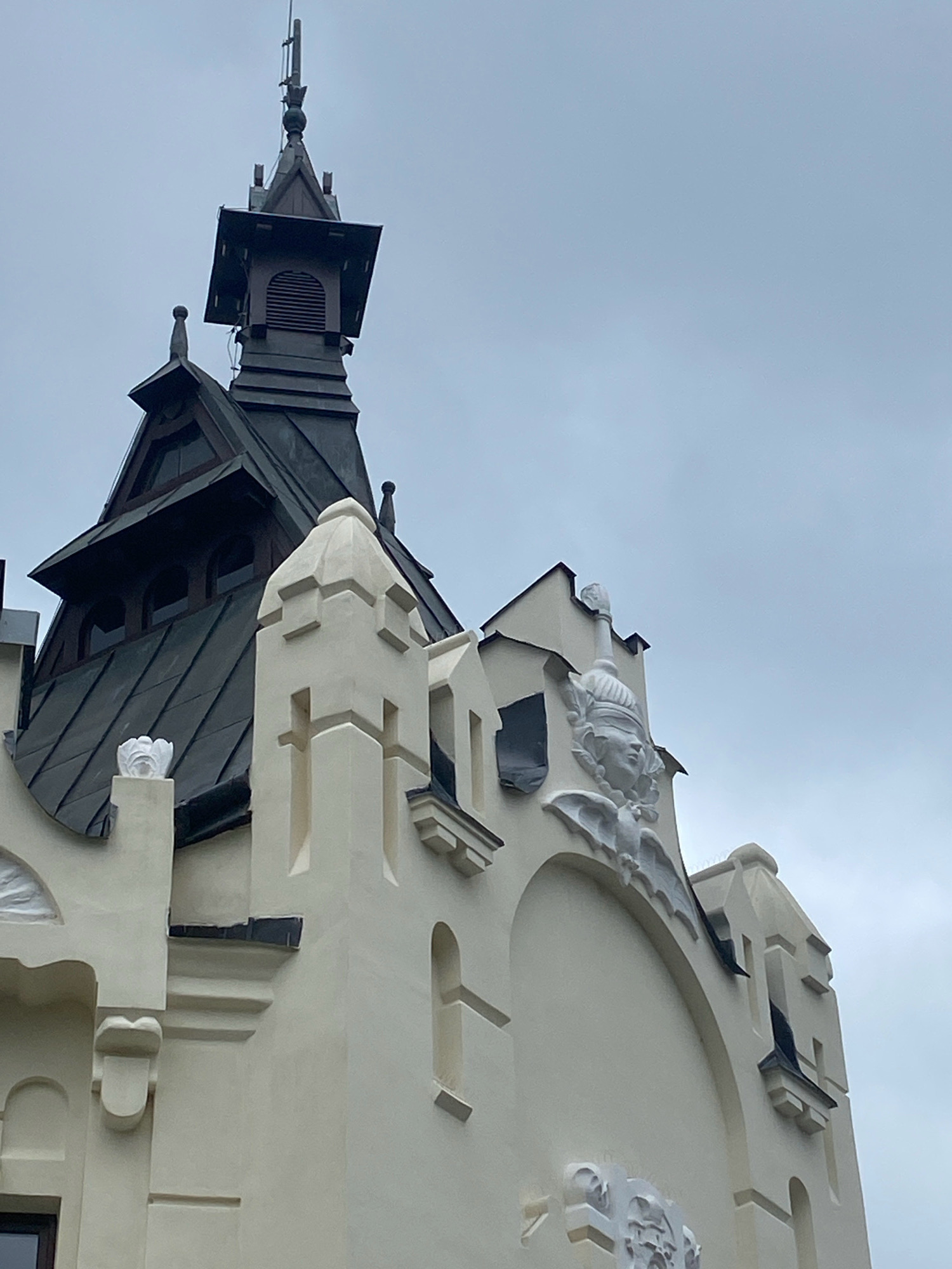 The image depicts a detailed view of the upper facade of a building, likely an architectural landmark, with a cloudy sky in the background. The building features ornate architectural elements, including decorative sculptures and a distinctive roofline.
On the left side of the image, a tall, dark-colored spire-like structure rises from the roof. This spire is topped with a thin antenna or lightning rod. Below the spire, a dark, multi-faceted roof section is visible, featuring arched windows. To the right of the spire, a lighter-colored, stepped architectural feature projects upwards. A small, white, sculpted element resembling a flower or shell adorns the top of a pillar on this section.
The central and right portions of the image showcase the building's facade, rendered in a light, creamy color. This section is characterized by its stepped gables and decorative sculptural elements. A prominent sculpture of a stylized face with a headdress is mounted above an arched section of the facade. Below this face, a winged creature, possibly a gargoyle or a symbolic representation, is sculpted. Further down, another decorative sculpted element is partially visible. The facade also includes small, dark-covered window openings and various decorative indentations and protrusions. The architectural style appears to be historical, possibly Art Nouveau or a similar eclectic movement, judging by the ornamentation and the form of the building.
The image is an exterior shot, taken from a low angle looking upwards towards the building. The lighting suggests an overcast day.
This architectural detail is from the Building of the former Savings Bank (Bank Handlowy) located at 1 Rynek Square in Nowy Sącz, Poland. The building was designed by architect Karol Kozłowski and built between 1910-1912.