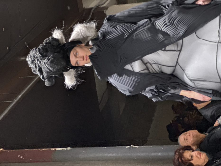 The image captures a model on a runway during a fashion show within an indoor venue in Paris, France, likely a repurposed industrial space or exhibition hall. The central figure, a model with fair skin and blue eyes, walks forward, gazing slightly to the viewer's right. They wear a prominent black and white sculptural headpiece featuring a large, textured black mass atop the head, flanked by white, fibrous protrusions, secured by a black cap. The model's attire consists of a light grey inner garment with visible black geometric stitching patterns across the torso and hips, covered by a voluminous, dark grey or black pleated outer garment with a structured, architectural form. In the lower-left foreground, several spectators are partially visible; two women are prominent, one with reddish-brown hair and another with very short light hair, both looking intently towards the model. Other indistinct figures are present in the dark background. To the far left, a vertical structural pillar with peeling paint, showing reddish-brown and lighter layers, marks the environmental context. The scene is brightly lit on the model, contrasting with the dark background.