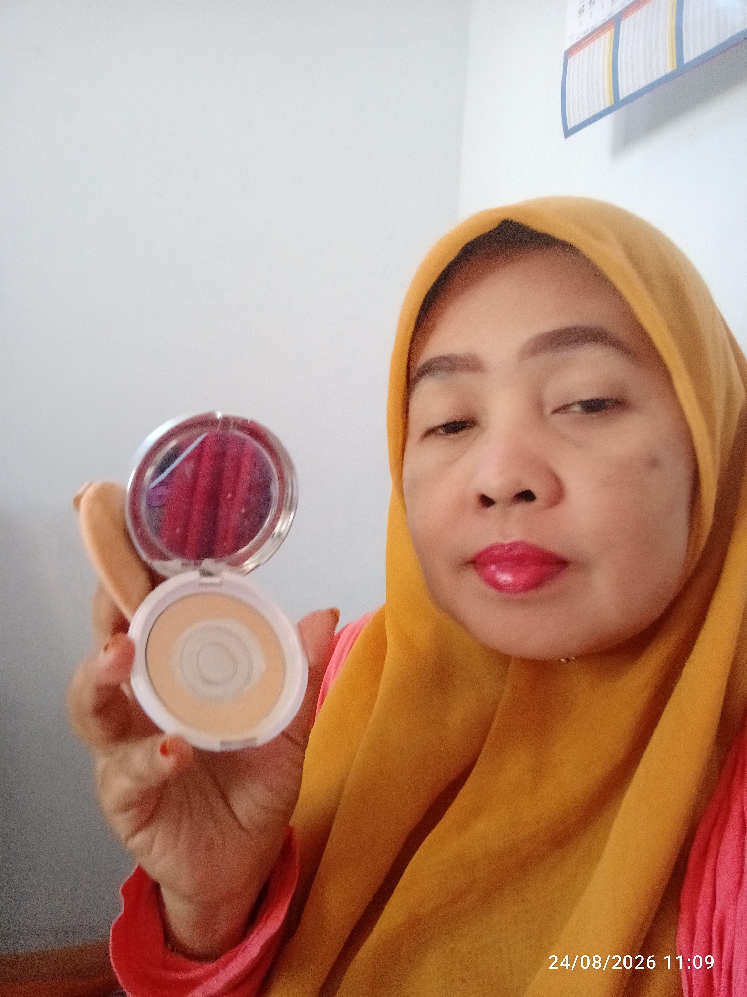 A woman wearing a yellow hijab and a pink shirt is holding an open compact powder with a puff in her left hand. She is looking at the camera, and her lips are painted with bright pink lipstick. The background is a plain white wall. The timestamp in the lower right corner indicates the image was taken on 24/08/2026 at 11:09. There is no visible indication of the location being Munjungan, Indonesia.