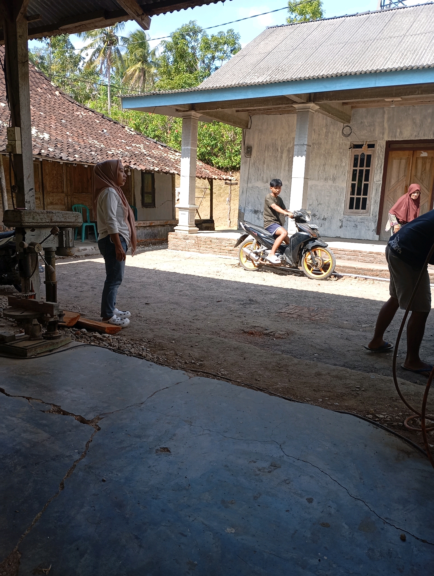 In Ketepung, Indonesia, a sunny outdoor scene unfolds in what appears to be a rural or semi-rural residential area. The foreground shows a cracked concrete floor. In the mid-ground, a woman wearing a light-colored shirt, blue jeans, and white sandals stands facing left, looking towards the right. She is wearing a light brown headscarf. To her right, a young boy in a dark t-shirt and blue shorts is sitting on a black motorcycle with yellow rims. Behind the boy and motorcycle is a simple concrete building with a corrugated metal roof and a wooden door. Further to the right, an older woman in a brown headscarf and patterned clothing is walking away from the building. In the background, traditional Indonesian houses with tiled roofs and wooden structures are visible, surrounded by lush green trees and palm trees under a clear blue sky. A piece of industrial equipment, possibly a drill press, is partially visible on the far left of the image.
