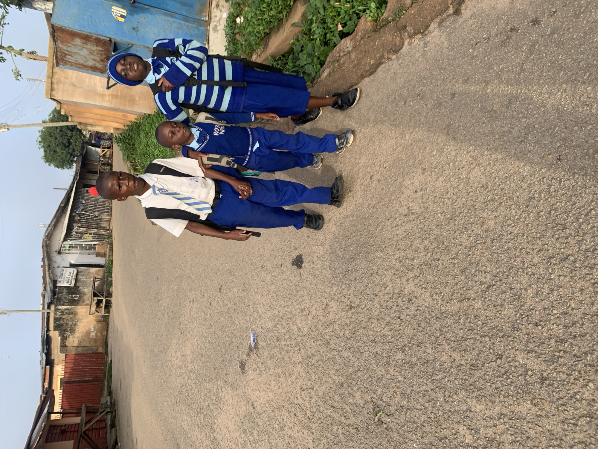 Three young children wearing school uniforms walk on a paved road in what appears to be a residential area of Jos, Nigeria. The child in the front, on the left, wears a white shirt, blue pants, a blue and white tie, and a black backpack. The middle child, slightly shorter, wears a dark blue uniform with a light blue tie and a dark backpack. The child on the right, a girl, wears a blue and white striped sweater over a blue uniform, blue skirt, and a black backpack. They are walking away from the camera and towards the background. The background shows a mix of buildings, some with corrugated metal roofs, and greenery. One building in the background has a sign that reads "TIM'S COLLECTION." The sky is overcast and light. The road is rough and appears to be made of asphalt with visible gravel. There is some trash on the road. The overall lighting suggests it is daytime.