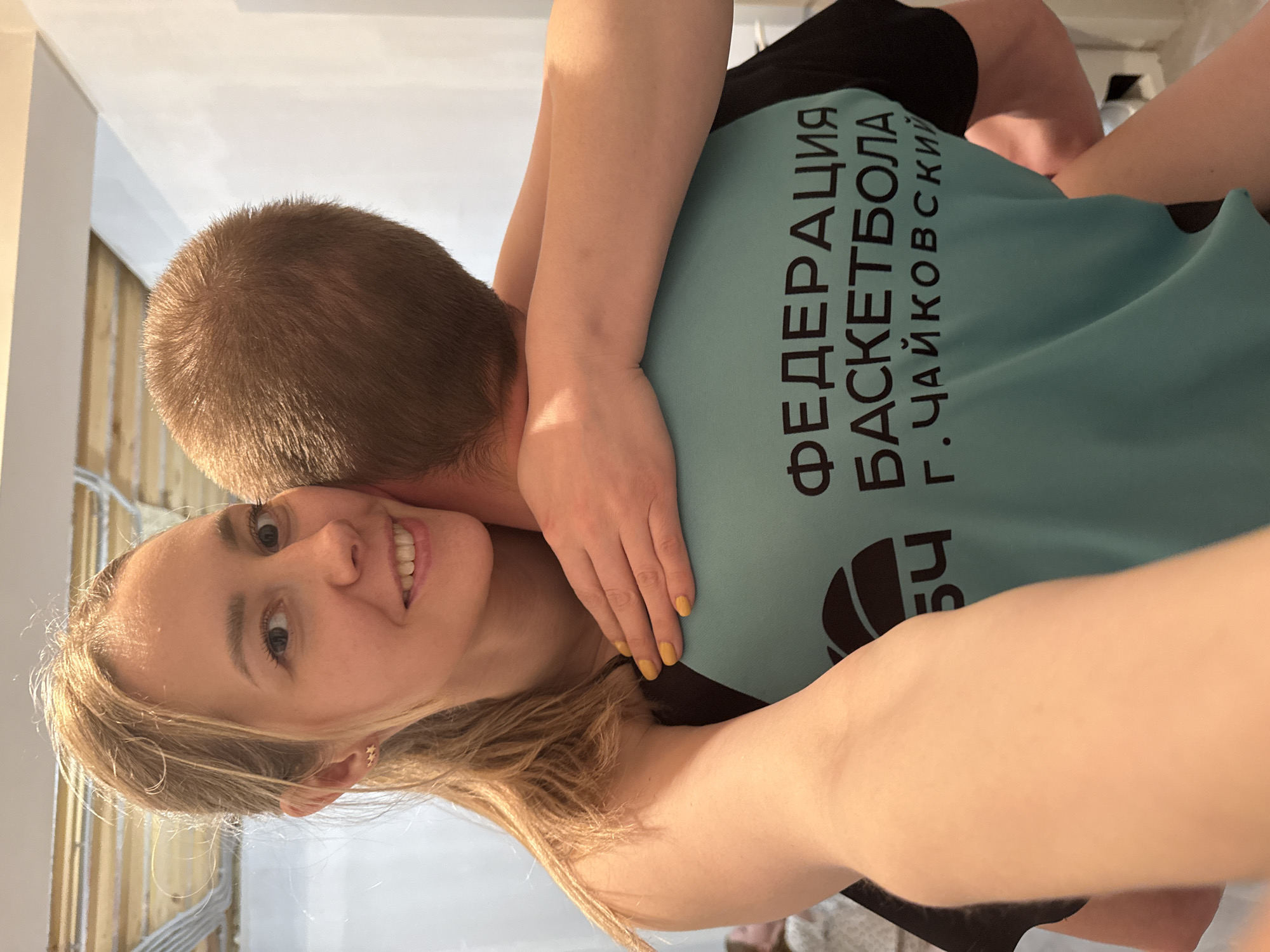 A woman with long blonde hair is smiling at the camera while hugging a man with short brown hair. The man is wearing a turquoise t-shirt with black sleeves and the words "ФЕДЕРАЦИЯ БАСКЕТБОЛА Г. ЧАЙКОВСКИЙ" in black Cyrillic letters. The woman's hand is visible on the man's neck, and her nails are painted yellow. The background is blurry, but it appears to be an indoor space with white walls and some wooden framing. The text on the shirt indicates the location is likely in the city of Chaikovsky, Russia.