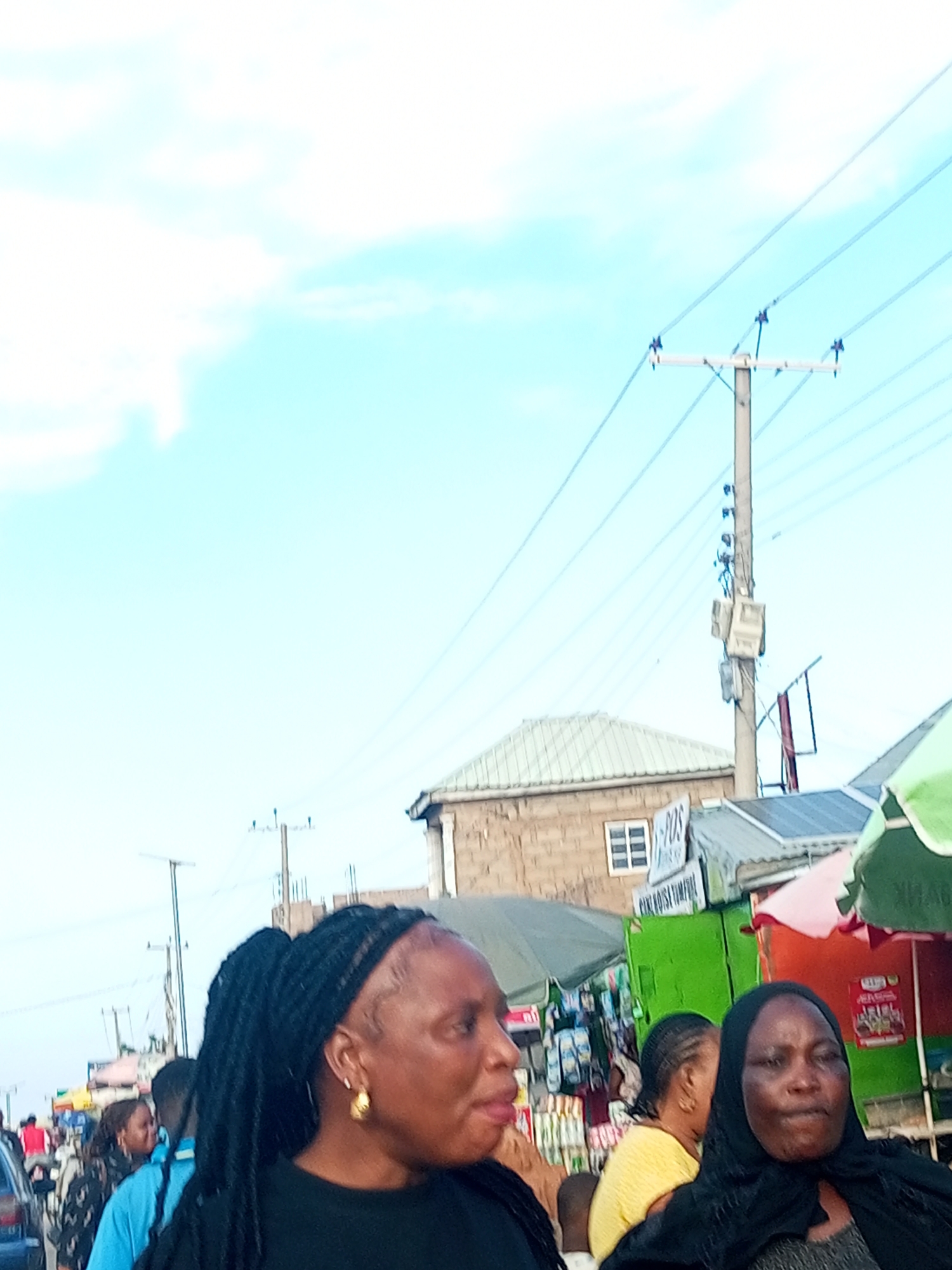 An outdoor street scene in Tempure, Nigeria, under a bright, cloudy sky. In the foreground, a woman with dark braided hair and gold earrings looks to her right. Behind her, other people are visible, some walking and others standing near market stalls. The stalls display various goods, and the overall impression is that of a busy marketplace. Electrical poles and power lines crisscross the sky, indicating an urban environment. A light brown brick building with a white-framed window is visible on the left side of the frame, suggesting a commercial or residential area. The scene appears to be set during daylight hours.