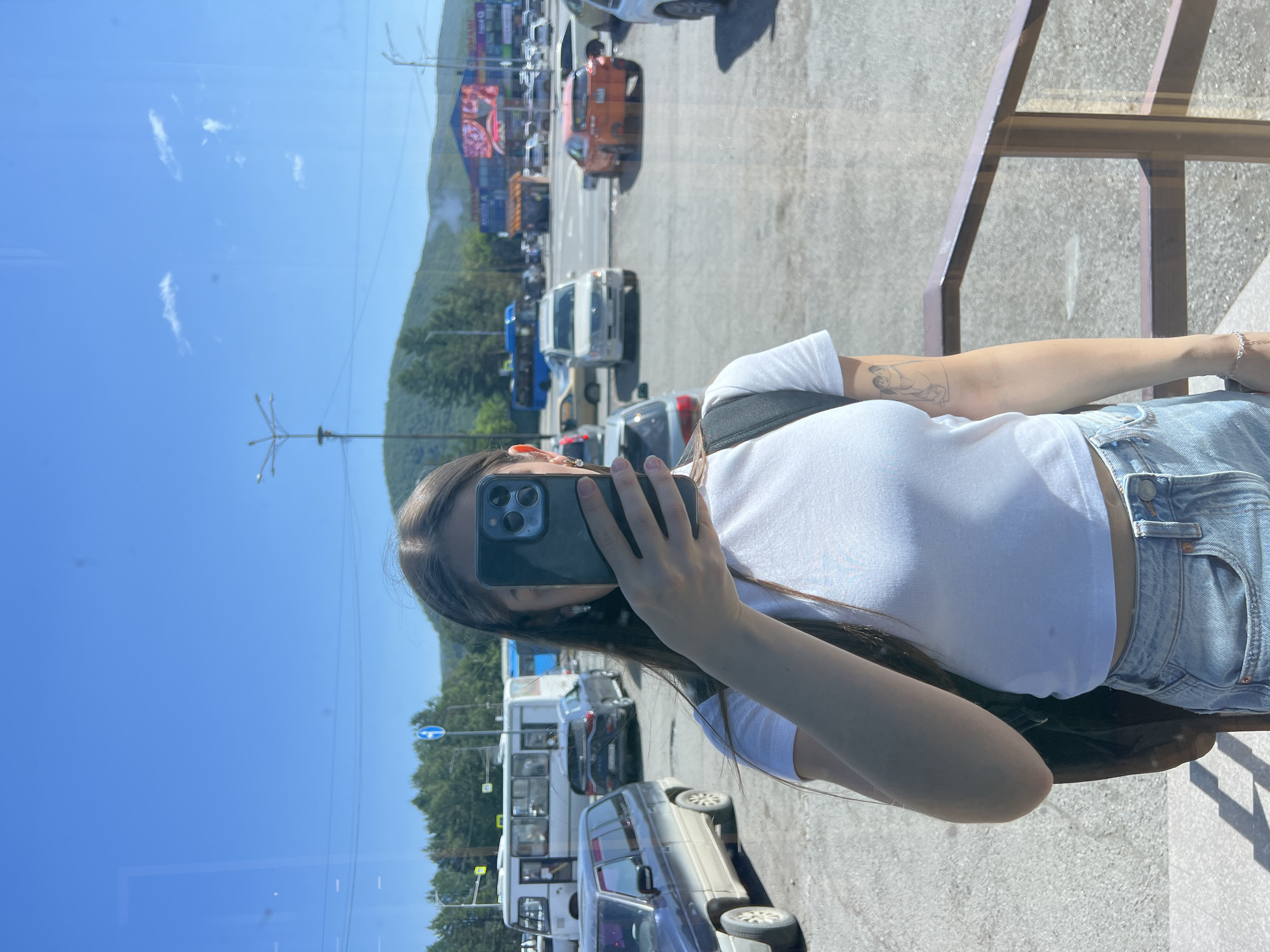 A young woman stands in front of a window, holding her phone to take a selfie. She is wearing a white t-shirt and denim shorts, and has long dark hair. Her left arm has a tattoo of two figures embracing. In the background, a parking lot is filled with various vehicles, including cars, buses, and a distinctive orange SUV. Beyond the parking lot, a town scene unfolds with buildings, trees, and hills under a clear blue sky. The environment suggests a sunny day in a town with a mountainous backdrop. Based on the presence of Cyrillic signage and the style of architecture, it is plausible this image was taken in Yelizovo, Russia, a town known for its proximity to volcanoes and natural landscapes.