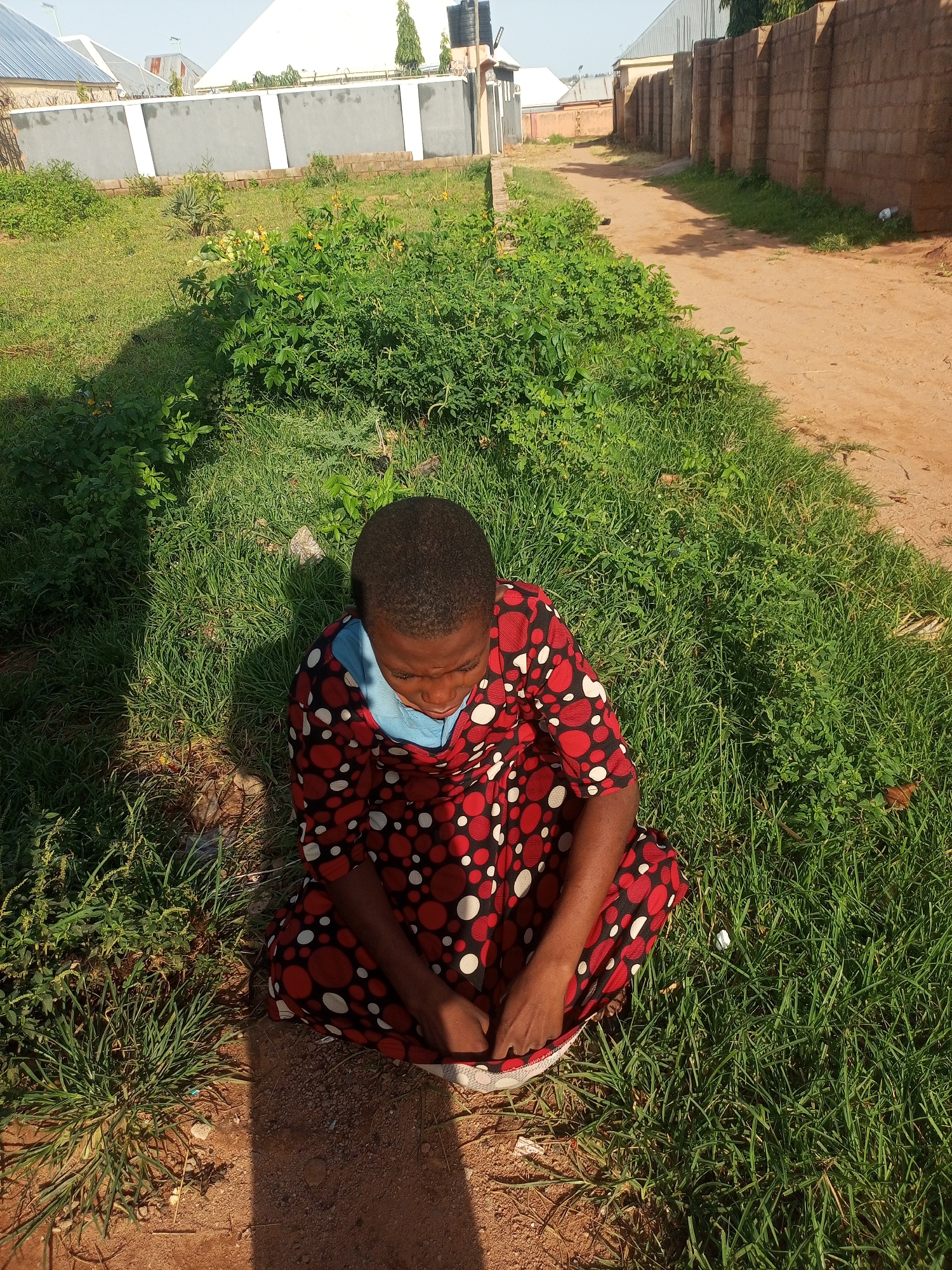 In Tempure, Nigeria, a young child sits cross-legged on a patch of green grass next to a dirt path. The child is wearing a red dress with a pattern of white and red circles, and a blue undershirt. The child is looking down and appears to be resting. The environment is outdoors, with a dirt road to the right and a grassy area to the left. In the background, there is a gray wall with white accents, and further back, buildings with corrugated metal roofs. The sun casts a shadow on the child and the grass.