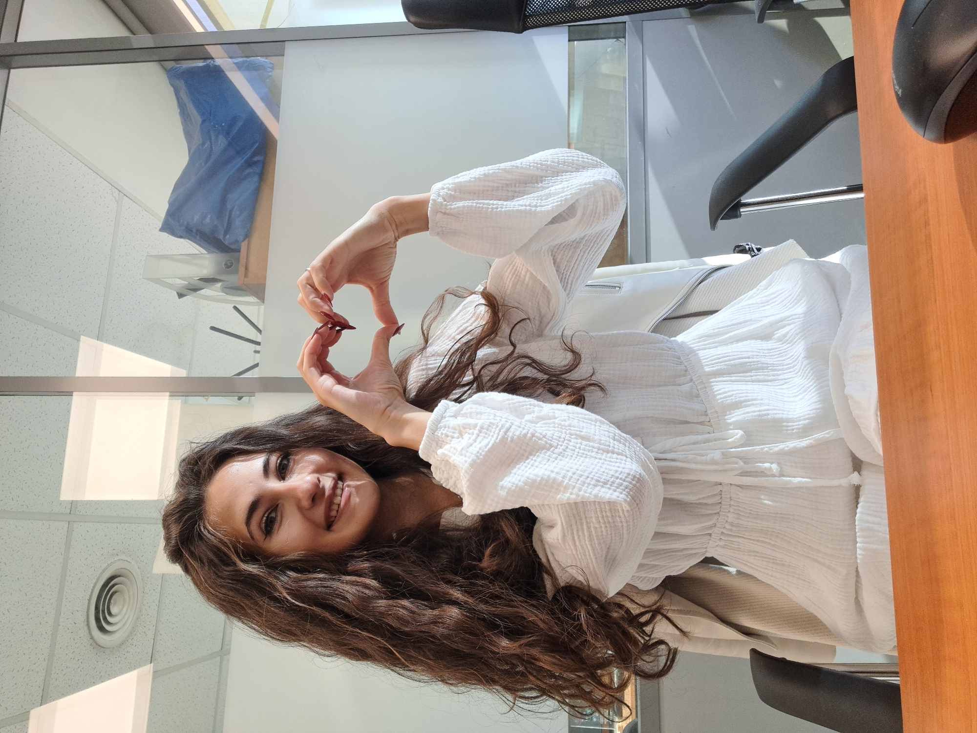 A young woman with long, curly brown hair is seated in an office chair, making a heart shape with her hands. She wears a white dress with long, puffy sleeves. The setting appears to be an office environment, with a ceiling vent visible, office dividers, a hanger on the chair and a wooden desk. The location is plausibly in an office in the city of Sofia, Bulgaria.