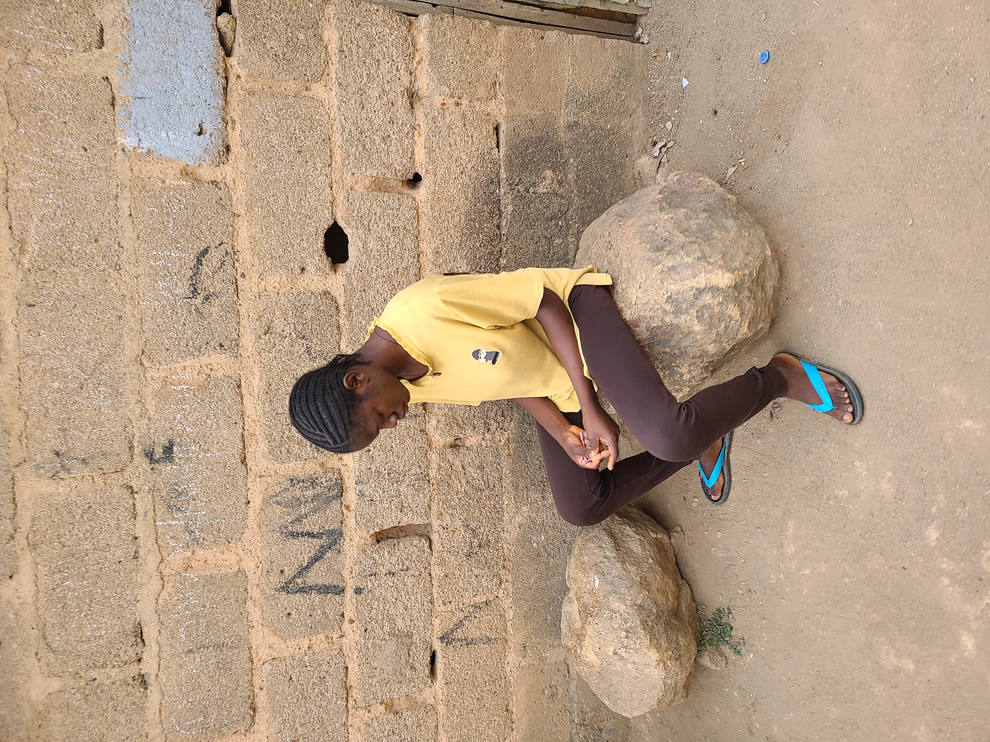 A young African woman in a yellow t-shirt and brown leggings kneels on a large rock outdoors in Jos, Nigeria. She wears blue flip-flops. Her head is close to a tan cinder block wall with grey paint. Another rock is near her feet. The ground is dirt.