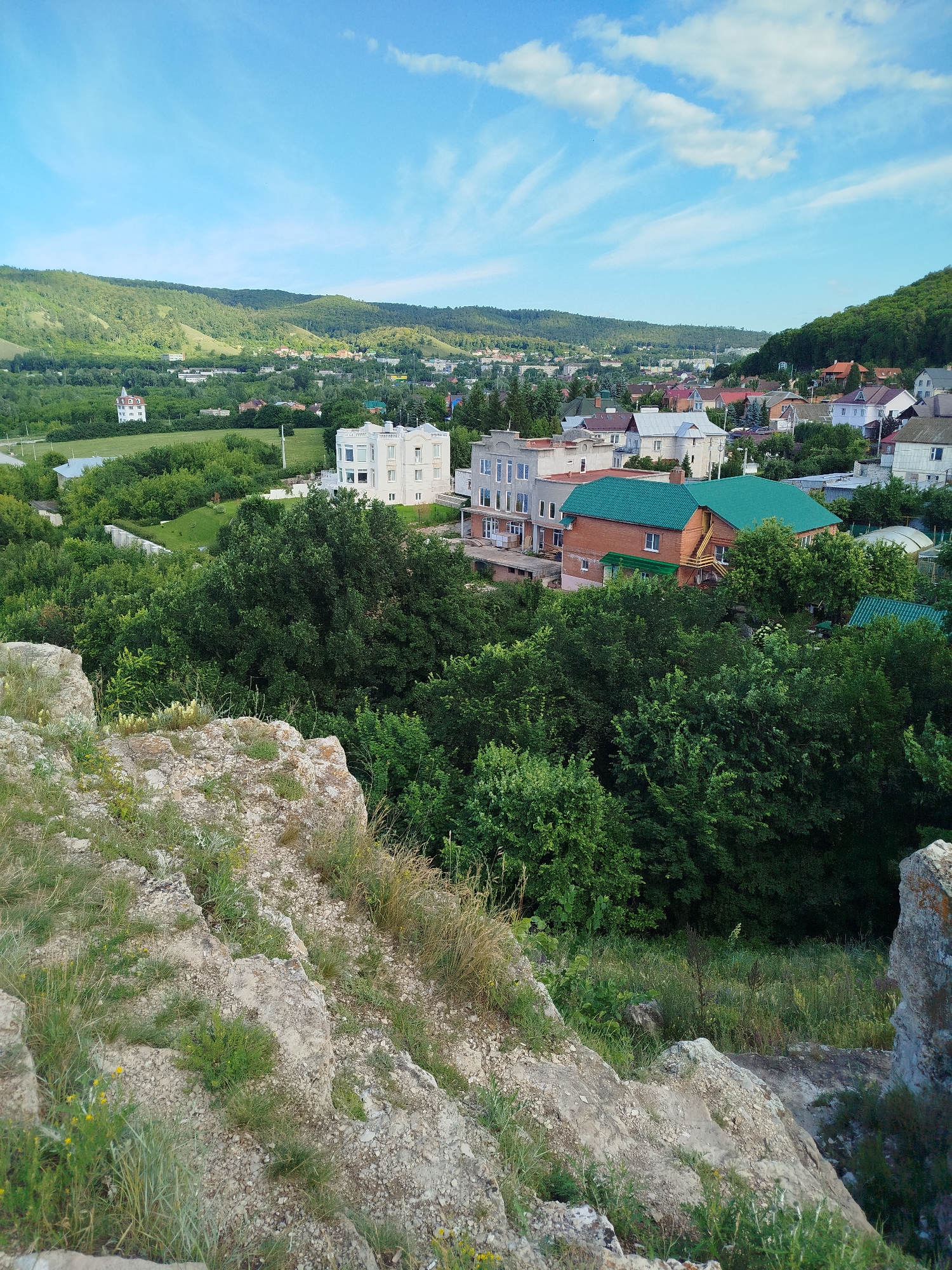 An outdoor, medium-shot, high-angle photograph captures a sunlit vista overlooking a town nestled within a valley. The foreground is dominated by a rough, rocky terrain covered in sparse grass and small yellow wildflowers, suggestive of a cliffside or elevated viewpoint. Beyond this rocky outcrop, a dense canopy of green trees stretches across the middle ground, gradually giving way to a suburban landscape in the distance.
The town features a variety of residential buildings with red and green tile roofs, alongside modern, multi-story structures with white and grey facades. A prominent white building with an ornate, classical-style facade stands out against the surrounding greenery in the mid-left of the frame. The town is situated at the base of gently sloping, verdant hills that rise in the background, their sides dotted with trees and some exposed patches of earth. The sky above is a vibrant blue, marked by wispy white clouds.
The scene is characteristic of the city of Zhigulyovsk, Russia, known for its picturesque location within the Samarskaya Luka National Park, which includes the Zhiguli Mountains. The topography visible in the photograph, with its combination of rocky outcrops, dense forests, and a settlement at the foot of hills, is consistent with the natural landscape of this region. The image depicts a tranquil, daytime scene, with no people visible. The overall impression is one of a scenic, semi-rural settlement in a mountainous environment.
