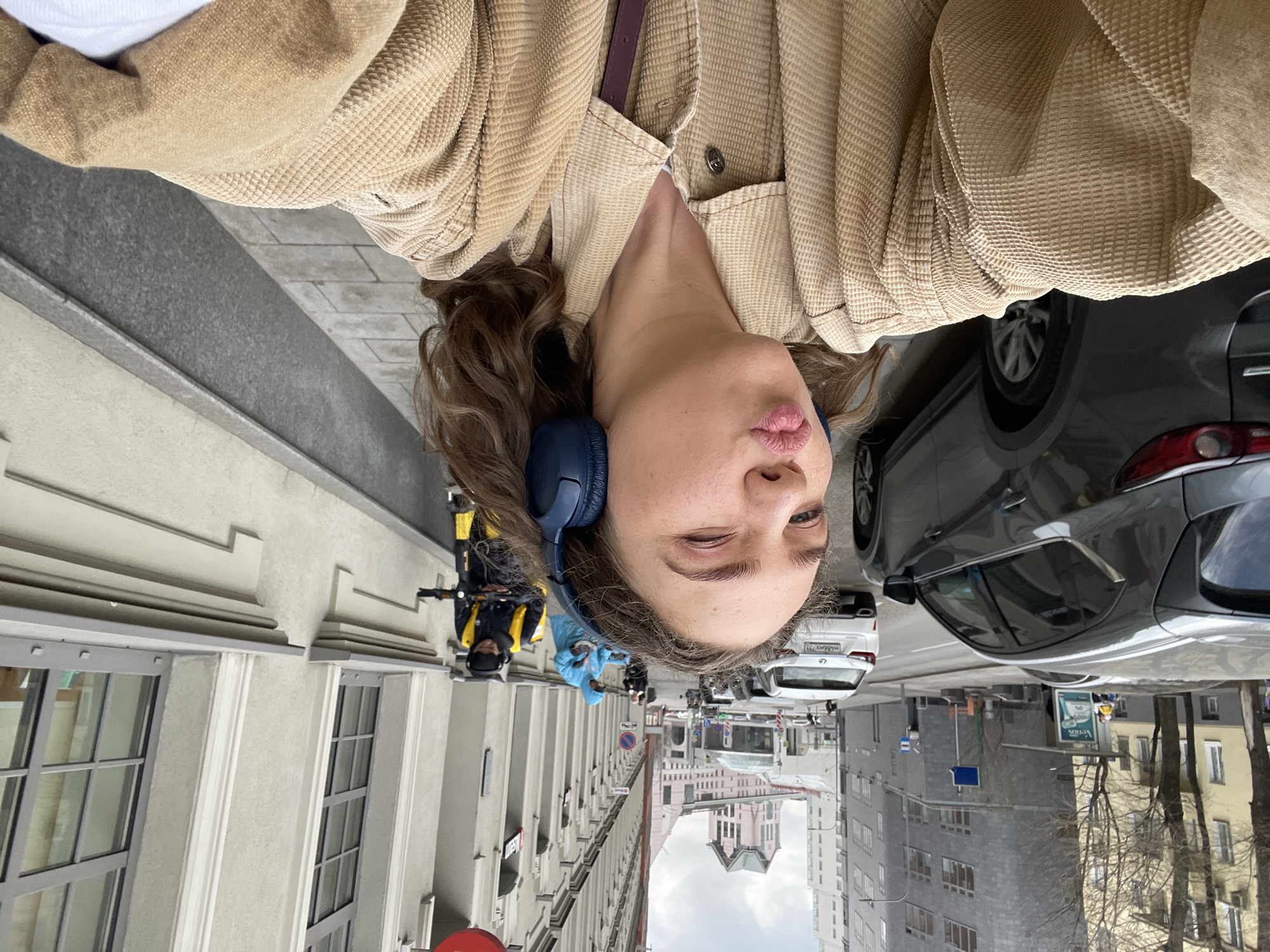 A woman with curly brown hair, wearing blue headphones, a beige corduroy jacket, and a white shirt, is taking a selfie. She has a slight smirk on her face. The background shows a city street. Buildings line the street on both sides, and a glimpse of the skyline is visible. Several cars are parked along the curb. Other people are visible further down the street, some wearing masks and riding scooters. This street scene is consistent with the city of Moscow, Russia.
