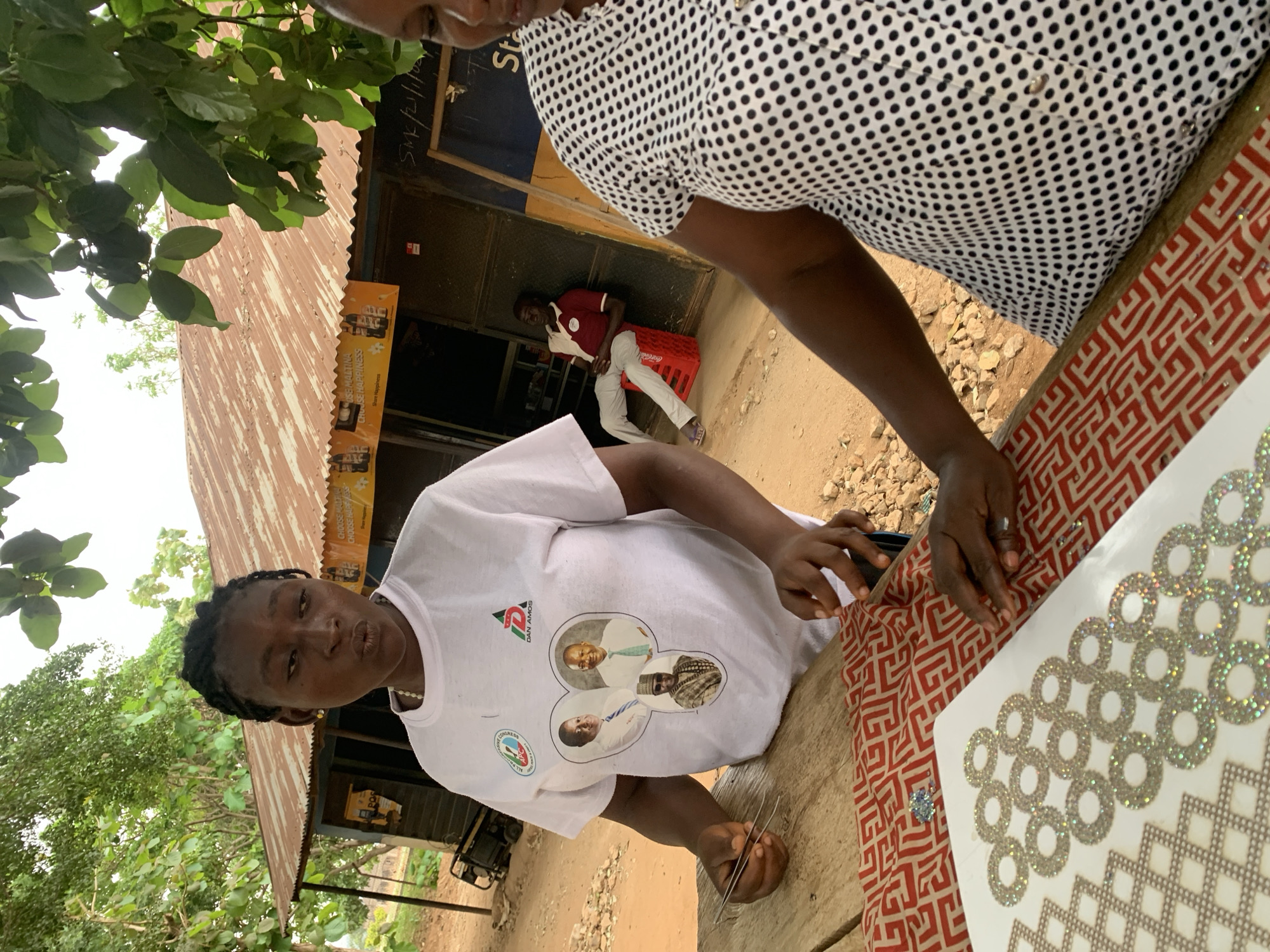 The image shows two individuals involved in a crafts-related task in Maigizo, Nigeria. One person, positioned behind a wooden table, is wearing a white shirt with black polka dots. The other individual, standing on the opposite side of the table, is wearing a white t-shirt emblazoned with a political figure and the text "DAN AMOS", along with a political logo. This person is working with a sheet of sparkling ring-shaped rhinestones, apparently adhering them to a reddish-brown fabric with a geometric pattern. She holds several slender metal sticks, possibly for manipulating the rhinestones.
In the background, a man is sitting on a red plastic crate, in front of a building with corrugated metal roofing and signs. A tree is partly visible on the left. The location appears to be a semi-outdoor workshop or a roadside business.