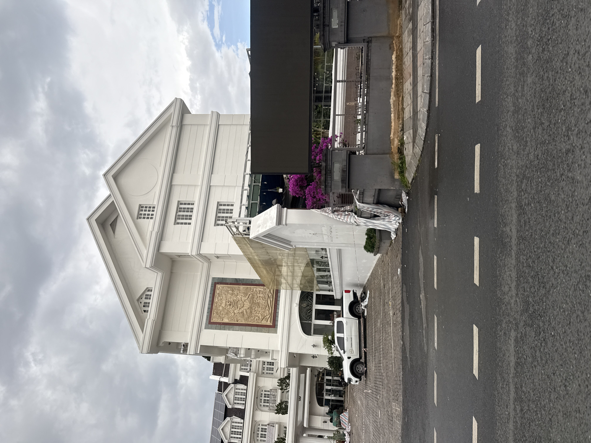 A white pickup truck with "THUHN PHAT" printed on its side is parked in a cobblestone lot in front of a large, modern white building with classical architectural elements. The building has multiple stories, white columns, and a large ornate relief on its facade. Purple bougainvillea flowers are visible near the entrance, and a large dark screen is mounted on the right side of the building. Construction materials, including striped tarps, are present in the foreground and near the building. The sky is overcast. The building is likely a hotel or resort, possibly located in the city of Da Lat, Vietnam, given the architectural style and landscaping often found in the region.