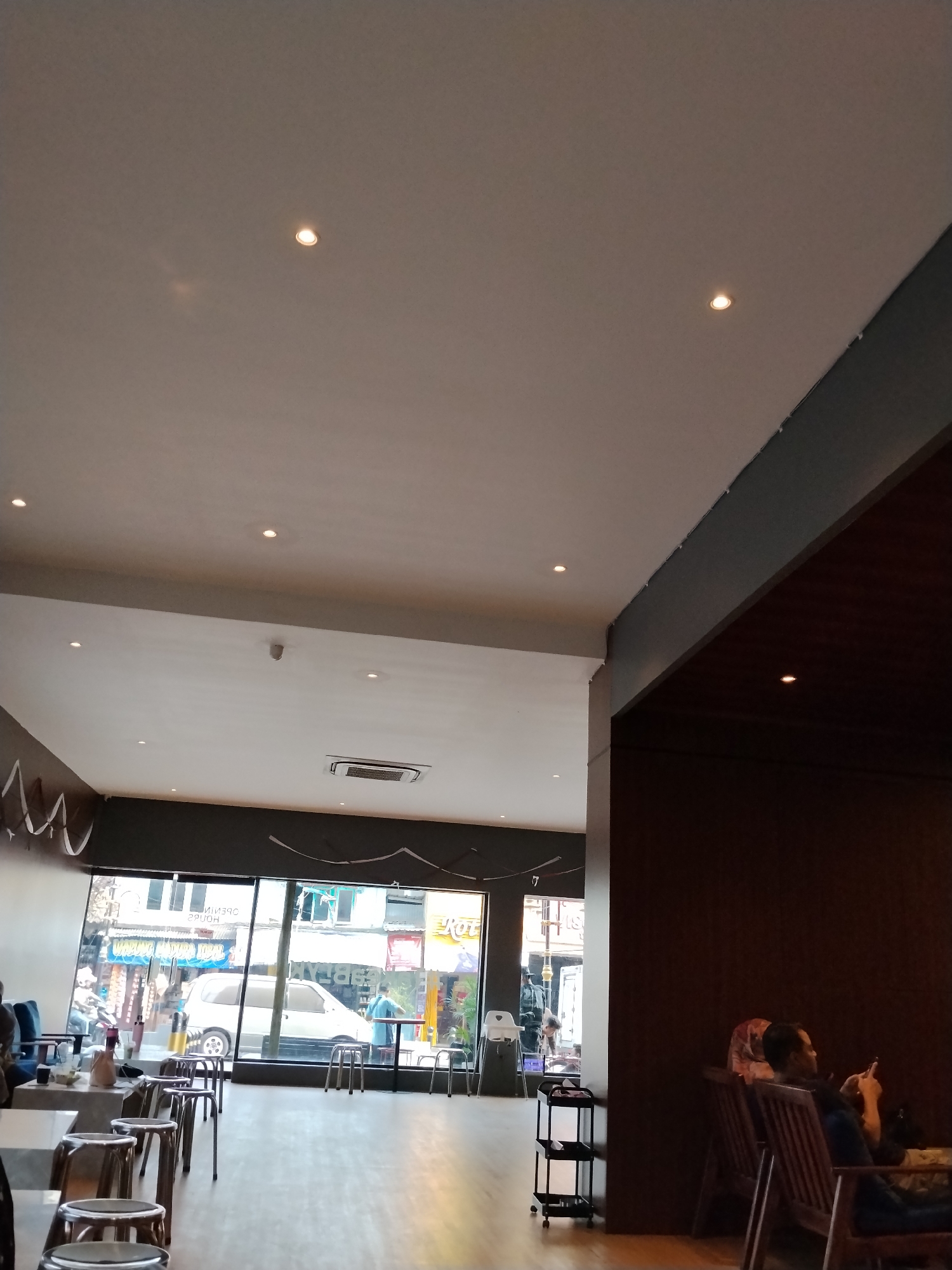 The image depicts the interior of a cafe in Serang, Indonesia, characterized by a modern, minimalist design. The ceiling features recessed lighting and an air conditioning unit. The walls are painted in neutral tones, with a darker accent wall in the foreground. The flooring is light-colored wood.
In the foreground, several metallic stools are arranged around low tables. One table appears to have some items on it, including a bottle. To the right, a person is seated in a wooden armchair, looking at their phone. They are wearing a headscarf.
Through a large glass window, the exterior of the cafe is visible, showing a street scene with buildings and a white van. People are seen inside and outside the window, suggesting an active commercial area.
The overall ambiance is casual and relaxed, suitable for a cafe setting. The lighting suggests it might be daytime or early evening.