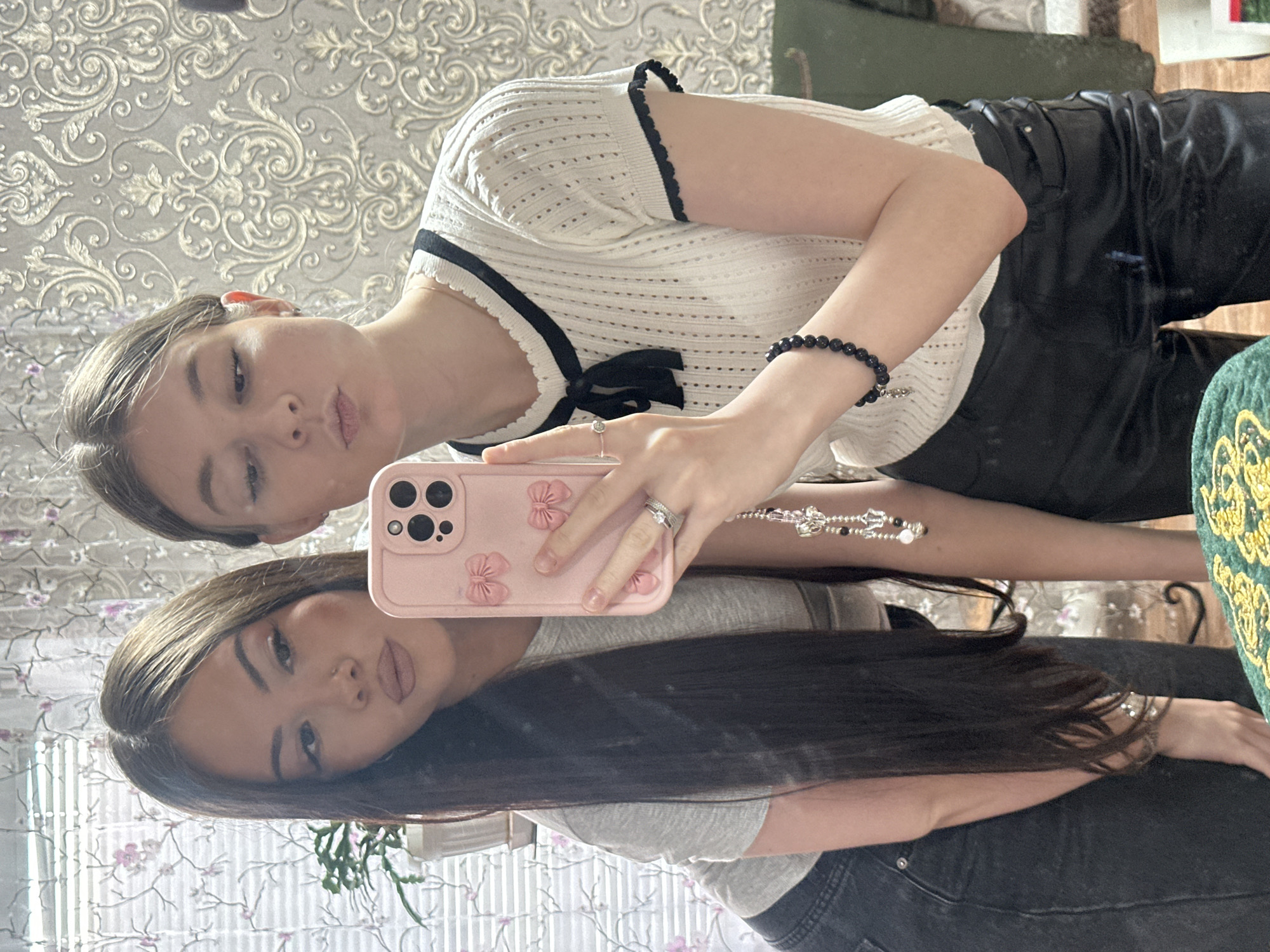 A mirror selfie featuring two women. The first woman, likely the subject, has light brown hair, fair skin, and is wearing a white short-sleeved knit top with black trim and a small black bow. She has a black beaded bracelet on her right wrist and rings on her right hand, which is holding a pink floral phone case. She is pouting at the camera. The second woman, her reflection in the mirror, has long dark hair, and is wearing a grey t-shirt. She is looking towards the camera. The background includes floral patterned wallpaper. The location is unknown, and there is no clear indication that it is in Коноваловка, Russia.