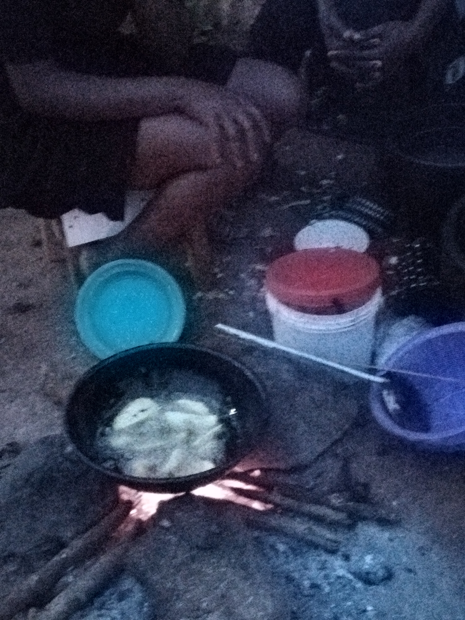In Du, Nigeria, an outdoor scene depicts individuals engaged in cooking. The central focus is a black frying pan placed over a fire, containing what appears to be fried food. To the left of the pan is a bright turquoise bowl. To the right, a white container with a red lid sits on the ground, accompanied by a purple bowl. The background is obscured by darkness, but hints of human legs and hands suggest the presence of at least two people actively participating in the cooking process. The ground is a rough, earthy surface. The lighting suggests it is evening or nighttime.