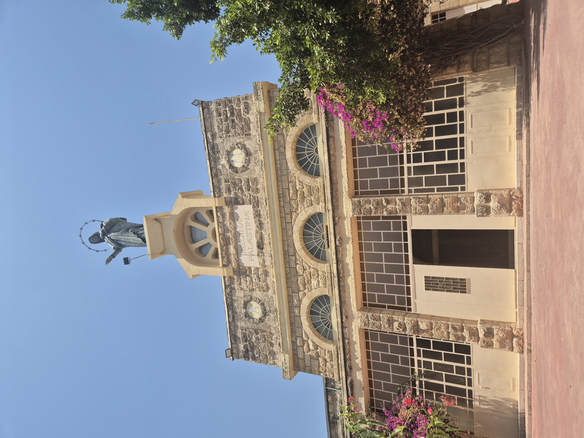 The image depicts the exterior of a building, identified as Regina Palestina in the Mate Yehuda Regional Council, Israel. The building features a stone facade with arched windows and a prominent statue of a robed figure with a halo atop the structure. The statue is positioned against a clear blue sky. Below the statue, a sign reads "REGINA PALESTINA". Large windows with a grid pattern and cream-colored doors are visible on the ground floor. To the right of the building, a lush green tree with visible branches and leaves is present, partially obscuring the structure. Pink and purple flowers are seen cascading from the tree and in front of the building on the left side. The ground in front of the building is a reddish-brown paved surface. The overall scene is bathed in bright sunlight, casting shadows on the building and ground.