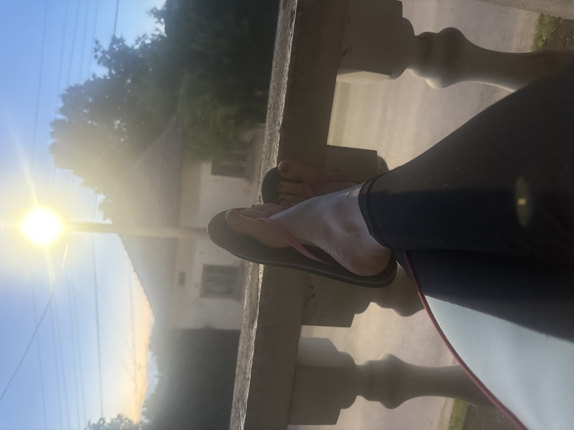 The image depicts an outdoor scene, likely captured from a balcony or porch overlooking a landscape in Iskar, Bulgaria. The perspective centers on the feet and lower legs of a person, dressed in dark leggings or pants, resting casually on a light-colored, ornate balustrade or railing. The person is wearing pink flip-flops, and red nail polish is visible on their toenails.
The sun is low in the sky on the left side of the frame, emitting a strong, bright light and creating noticeable lens flare, suggesting either sunrise or sunset. The clear sky transitions from a vibrant blue at the top to a warmer, lighter tone closer to the horizon. In the background, indistinct houses or buildings with pale facades are visible, partially obscured by trees and foliage. Overhead, a few power lines stretch across the sky.
The scene conveys a sense of peace and relaxation, as if the person is enjoying a quiet moment, observing the end or beginning of a day in a serene, possibly rural or semi-rural, environment. No discernible text is visible in the image.