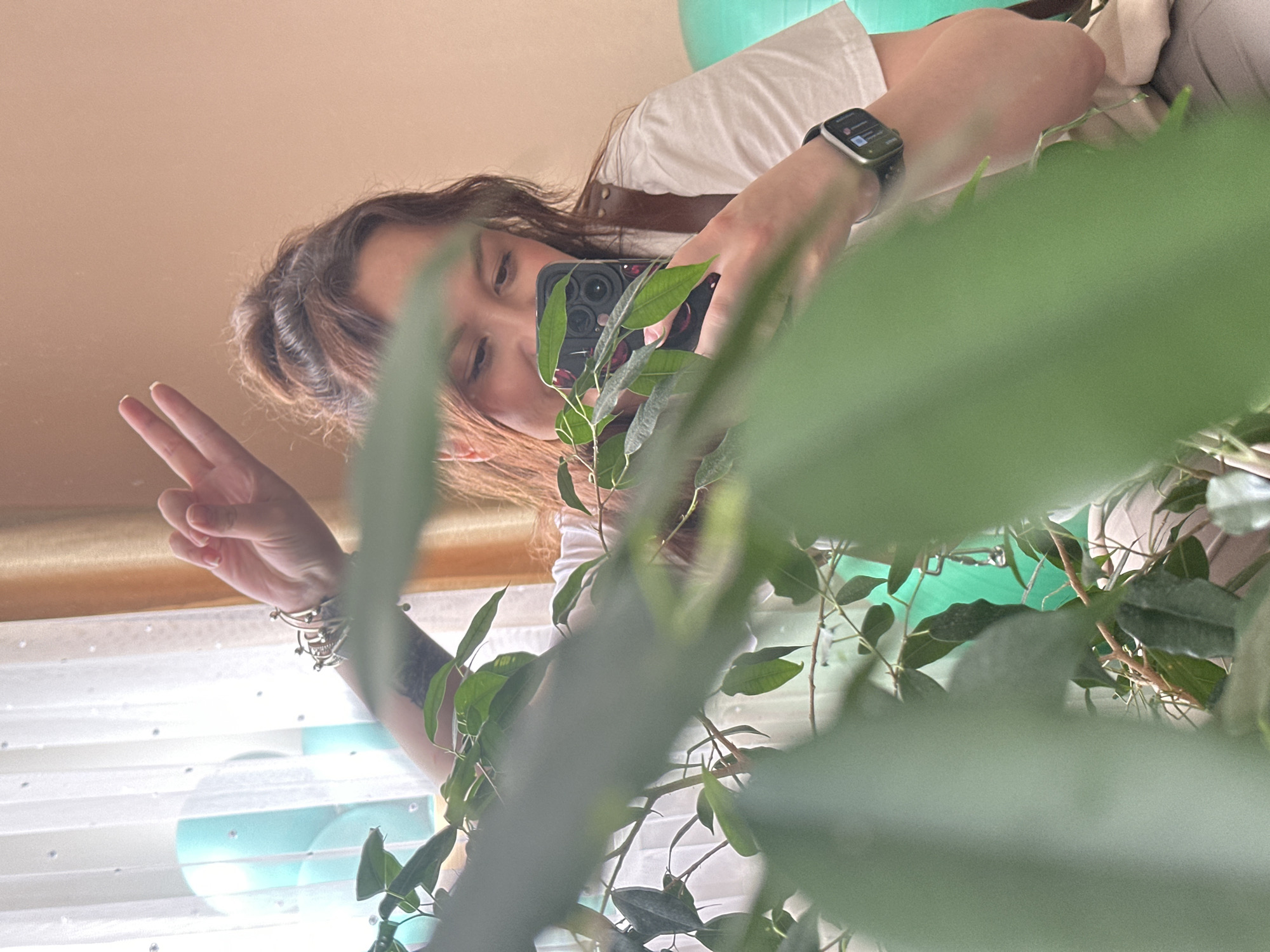 A person is taking a selfie with their smartphone. The person is wearing a white shirt and an Apple watch. They have one hand up in a peace sign and several bracelets on their wrist. A plant is obstructing a clear view of the person. The location appears to be indoors.