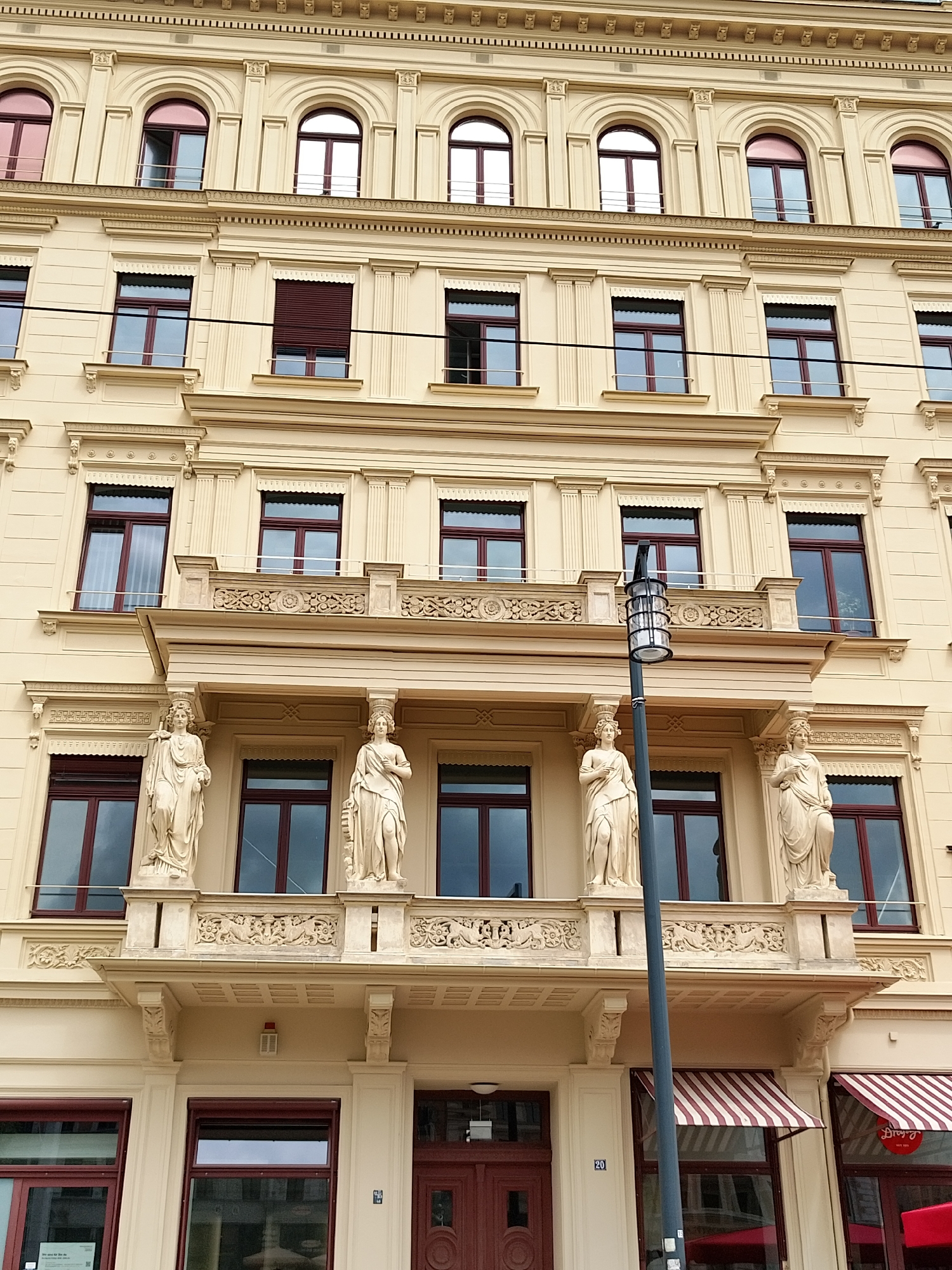 The image showcases the facade of a classical building in an urban environment. The building, likely located in Europe due to its architectural style, features several stories with multiple windows, some of which are arched on the top floor. The facade is primarily beige, adorned with decorative elements such as columns, cornices, and sculpted figures on a balcony. The windows are framed in a dark color, possibly dark red or brown. A lamp post is visible in the foreground, slightly obscuring the view of the building's central section. On the ground level, there is a store front with a red and white striped awning. The overall impression is that of a well-maintained, historical building in an urban setting.