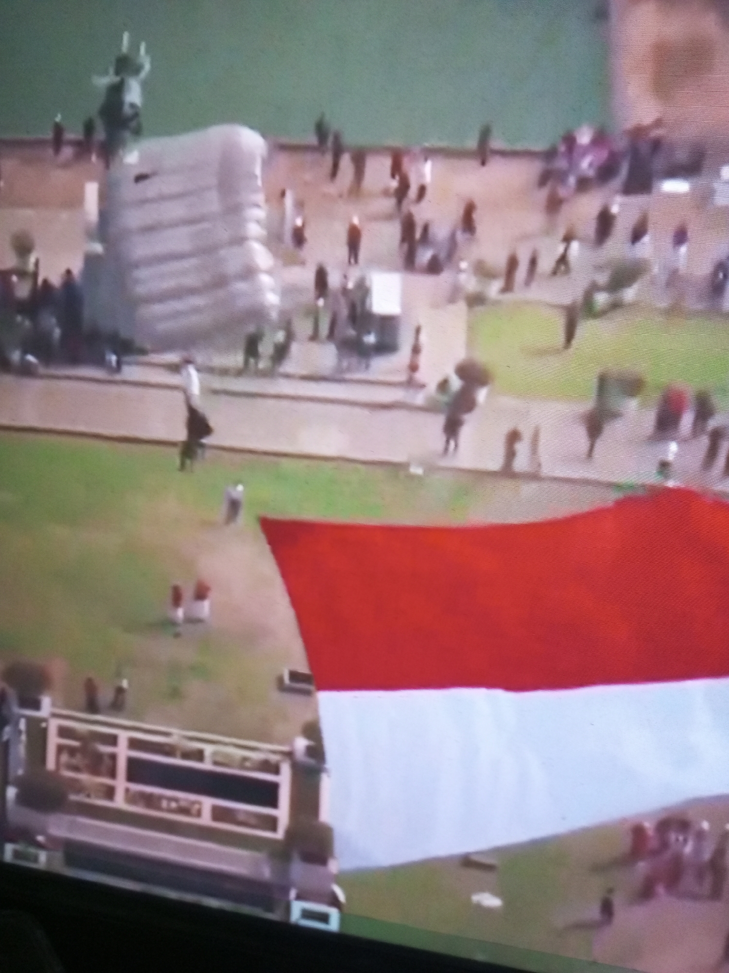 In Banyudono, Indonesia, an aerial view captures a large inflatable structure resembling a camel, adorned with horns and a hump. The camel is positioned on a light-colored surface adjacent to a green grassy area, where numerous people are congregated. In the foreground, a large Indonesian flag, red and white, is prominently displayed, partially obscuring the view of the scene. A long, white barrier with dark rectangular panels runs across the bottom of the image.
