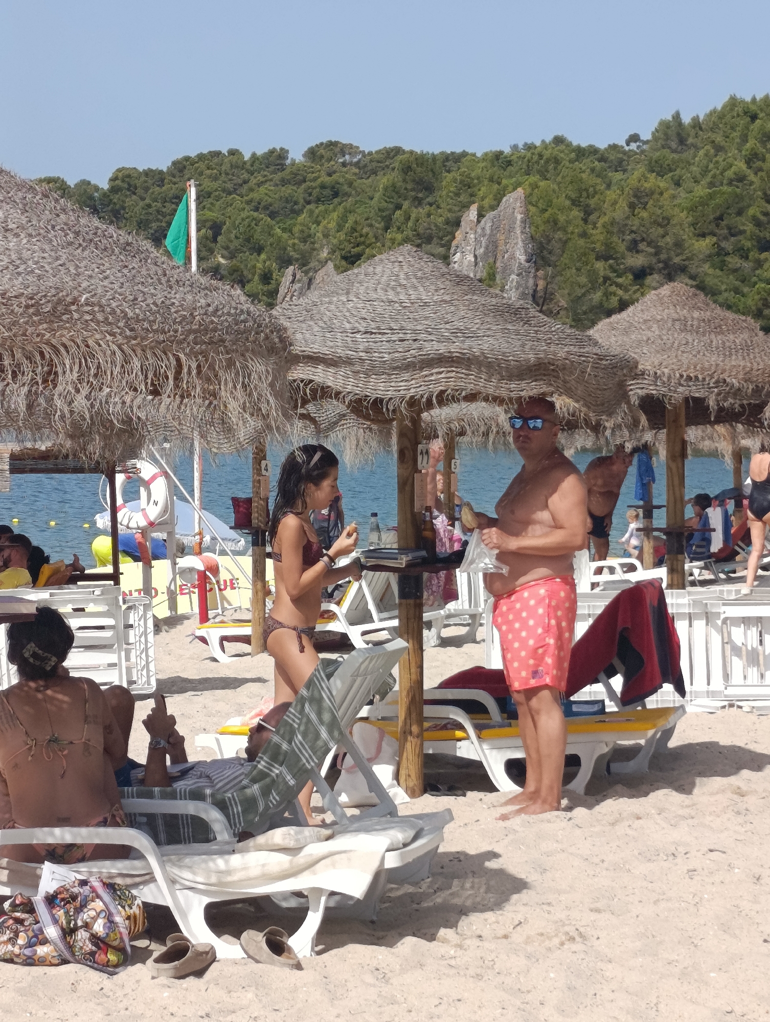 This image depicts a crowded beach scene under a bright, clear sky, with lush green trees and rocky outcrops visible in the background. The foreground is dominated by a sandy beach scattered with several thatched umbrellas and empty white lounge chairs. A group of people are present, some relaxing on lounge chairs, others standing.
In the mid-ground, a young woman in a patterned bikini is standing near a thatched umbrella, holding an object in her hands. To her right, a man wearing pink polka-dot swim trunks and sunglasses is also standing, holding a piece of food or a small item in his hand. He appears to be interacting with the young woman, possibly handing her something or engaging in conversation.
Further in the background, more beachgoers are visible, some obscured by umbrellas and lounge chairs. A rescue buoy with a red stripe and the letter 'N' is hung on a post near the water's edge, suggesting a lifeguard station or a designated swimming area. The sea is a calm blue, with a few buoys floating in the distance. The overall impression is that of a lively summer day at a popular beach resort. It is not possible to definitively determine if this location is in Setúbal, Portugal, based solely on the visual information provided.