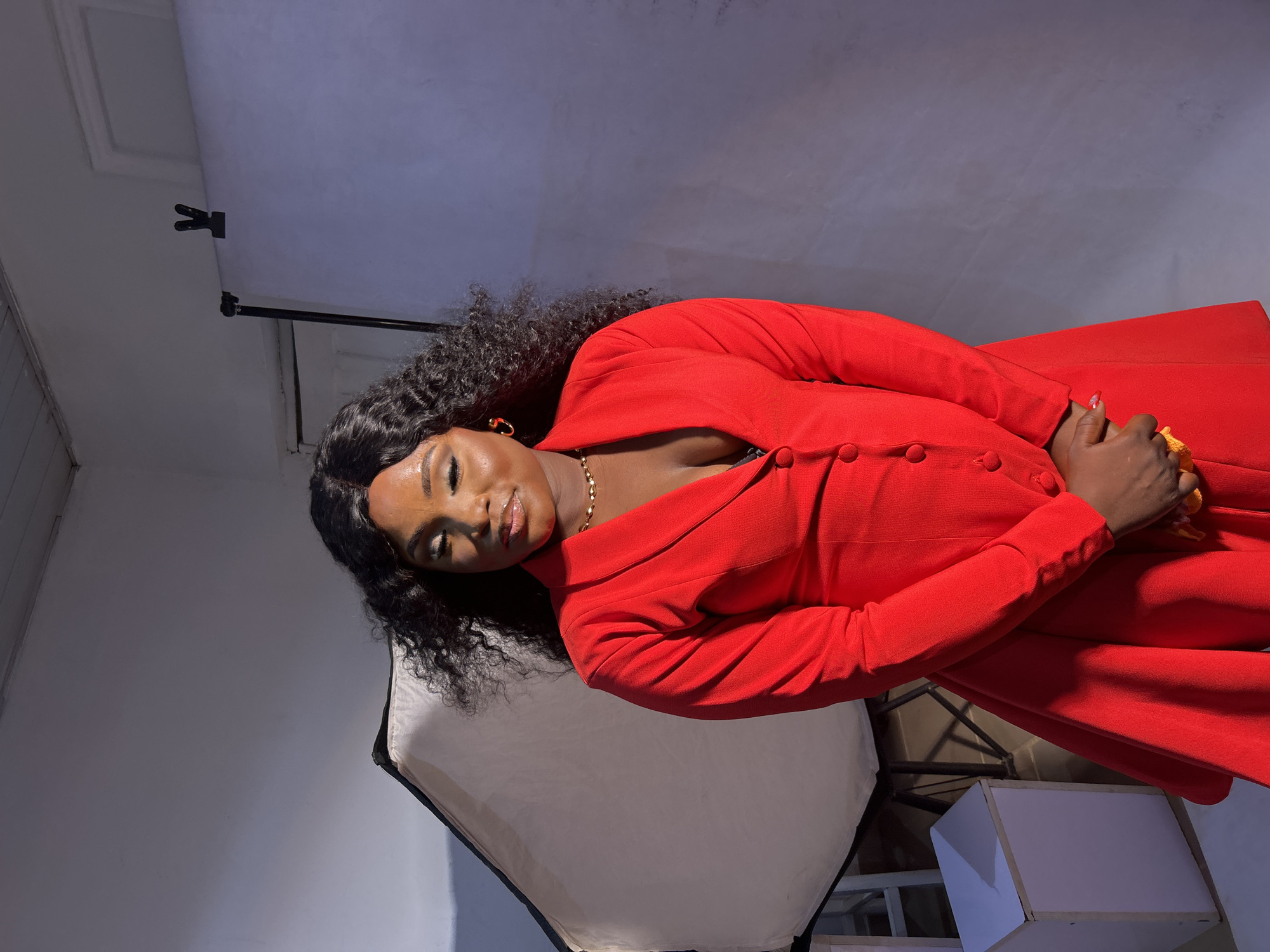 A young woman with dark hair is posing in a studio. She is wearing a red blazer. There is a softbox to the left of the woman, and a lighting setup behind her. The background appears to be a painted wall. It is unlikely this photo was taken in the city of Jos, Nigeria, as studio environments are not geographically dependent.