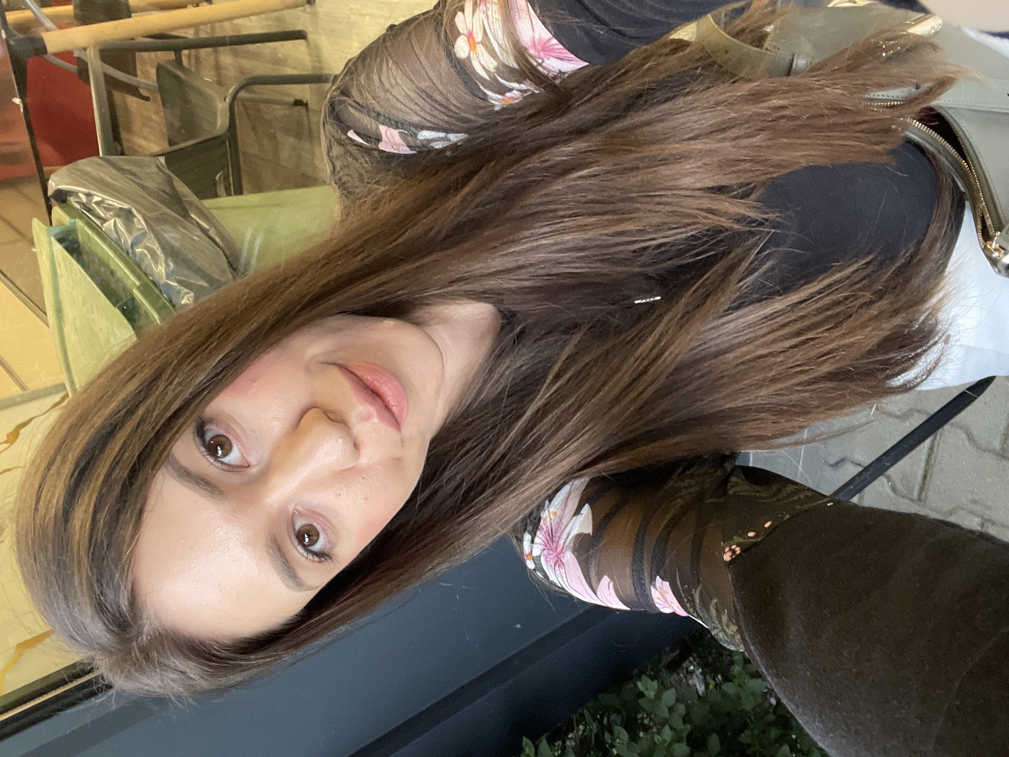 A woman is taking a selfie outdoors. The woman has long, brown hair and light skin. She is wearing a black top with floral patterned sleeves. She has brown eyes and pink lipstick. Behind her, there are tables and chairs, some covered with a plastic tarp, and some green foliage. The background suggests a patio or outdoor seating area. Based on visual analysis, the location is not immediately discernible as Sofia, Bulgaria without additional context or information. The image provides no specific clues that tie it to this city.