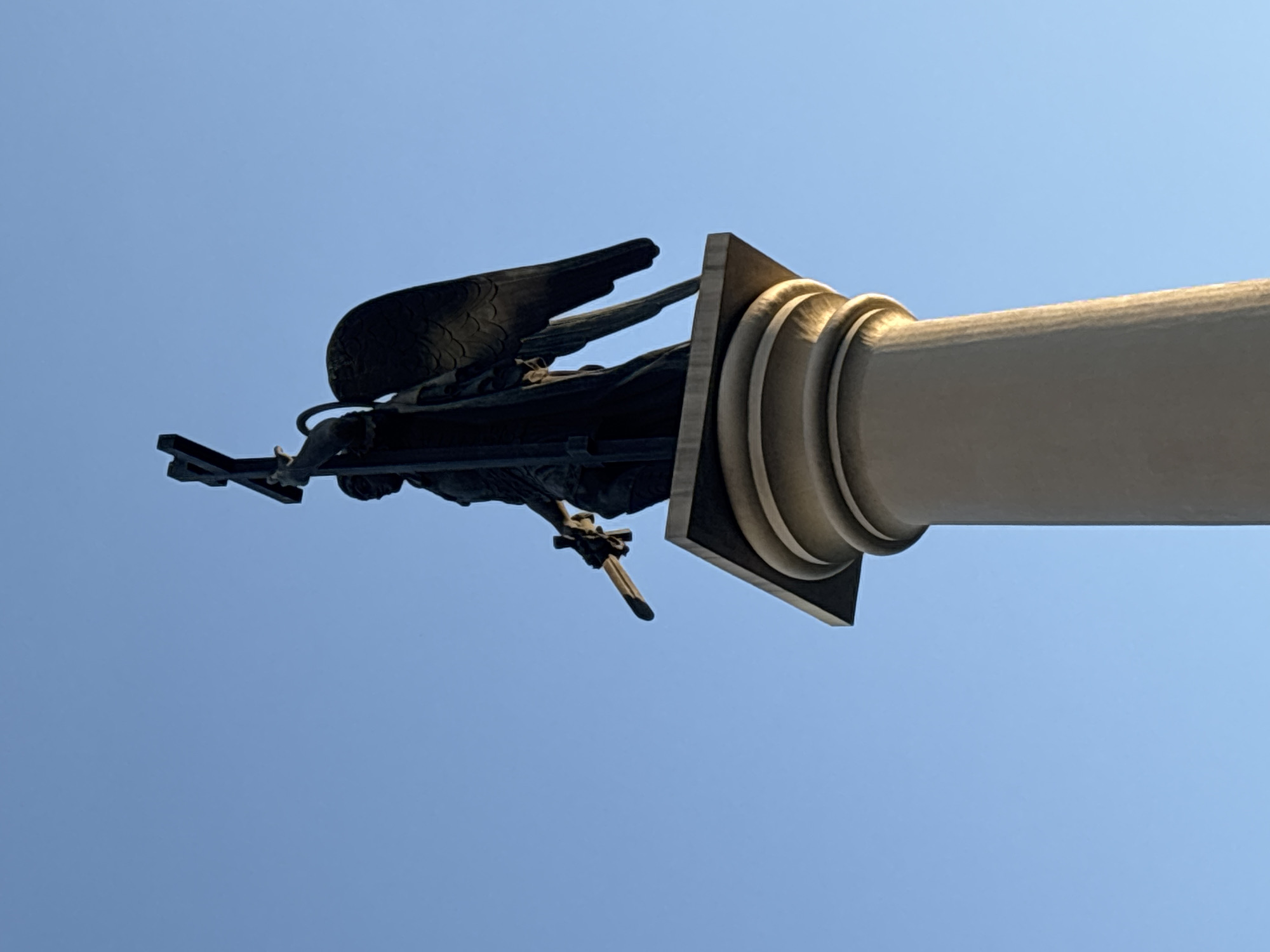 This media file shows a close-up, low-angle view of a bronze statue of a winged figure, likely an angel or a personification of victory, holding a sword. The statue is mounted on top of a fluted column. The background is a clear, bright blue sky, suggesting fair weather and daytime. The lighting on the column indicates it is illuminated by sunlight from the side.
The statue itself is dark bronze, with detailed wings spread outwards and a human-like form. The figure's right arm is extended, holding a sword with its blade pointing downwards. The column is a light, neutral color, possibly stone or painted concrete, and features classic architectural fluting. The statue and column are positioned against a vast expanse of sky, creating a sense of grandeur and elevation.
The image is cropped tightly, focusing on the upper portion of the column and the statue, with no ground or surrounding environment visible. This framing emphasizes the scale and artistry of the monument. The image was captured in Sochi, Russia. No text is visible in the image. The overall impression is one of solemnity and strength, often associated with monuments of this nature.