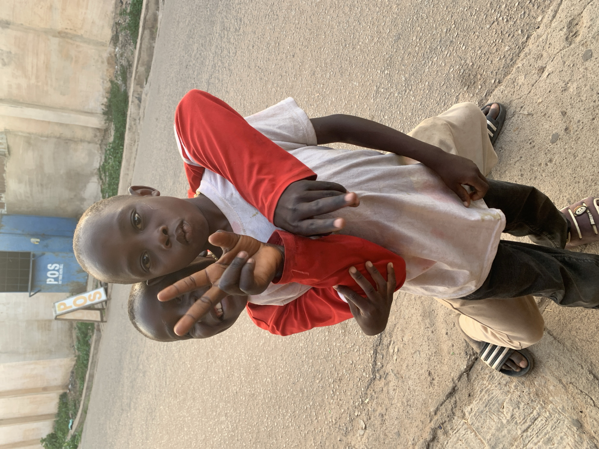 Two young Black African boys are outdoors on a concrete or asphalt surface next to a street. One boy wears a long-sleeved red and white shirt, tan pants, and Adidas sandals. The other boy is behind him. The boy in front is making a peace sign with his fingers toward the camera. Behind them, a blue structure with the letters "POS" visible on a sign. This suggests the scene is located in Jos, Nigeria.