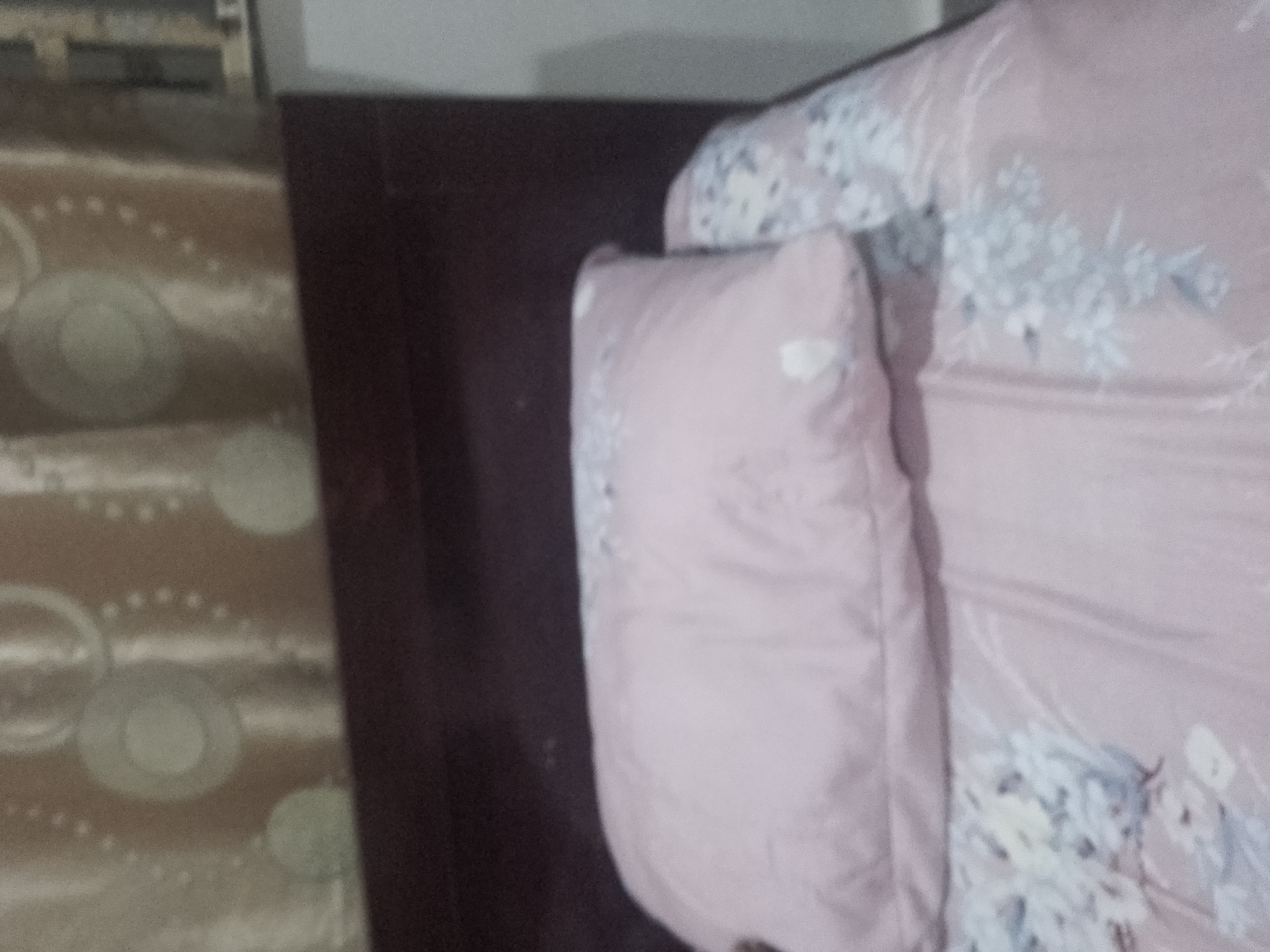 The image shows a close-up, somewhat blurry, view of a bed with pink floral patterned bedding and a pink pillow. The pillow is in the foreground, and part of the bedding is visible behind it, showing a delicate floral design in light blue and white. To the left of the bed, a patterned surface with circular designs is visible, possibly a rug or carpet. The background is a dark, solid surface, likely the headboard or wall of a room. The lighting is dim.