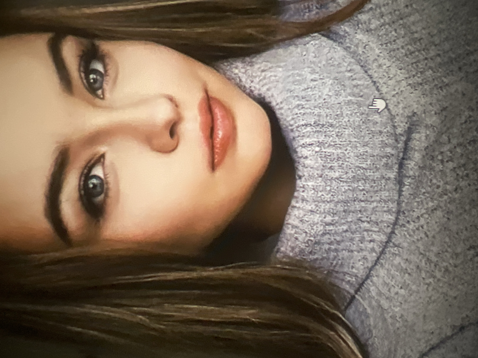 The image is a close-up portrait of a young woman wearing a light purple turtleneck sweater. Her face is the primary focus, with her eyes looking directly at the camera. She has light brown hair that falls around her shoulders. Her makeup includes eyeliner and mascara on her eyes and a glossy pink lipstick on her lips. The background is out of focus, but appears to be indoors. There is no indication of the city of Qarağandı or its administration in the image.