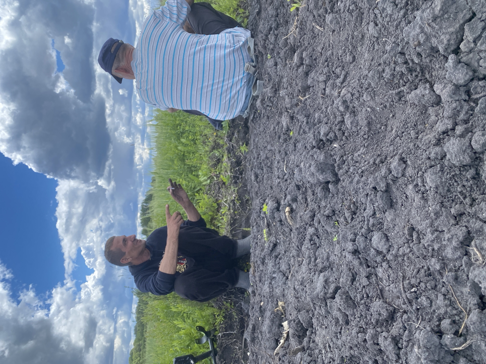 Two men are in a cultivated field near Pidluky, Ukraine. One man, wearing a striped shirt and cap, sits on the edge of the field. The other man squats in the field, holding an object, possibly a cigarette, in his hand. A metal detector is visible near the squatting man. The sky is visible with a few clouds, behind the men there is green grass. The field is dark and freshly turned.