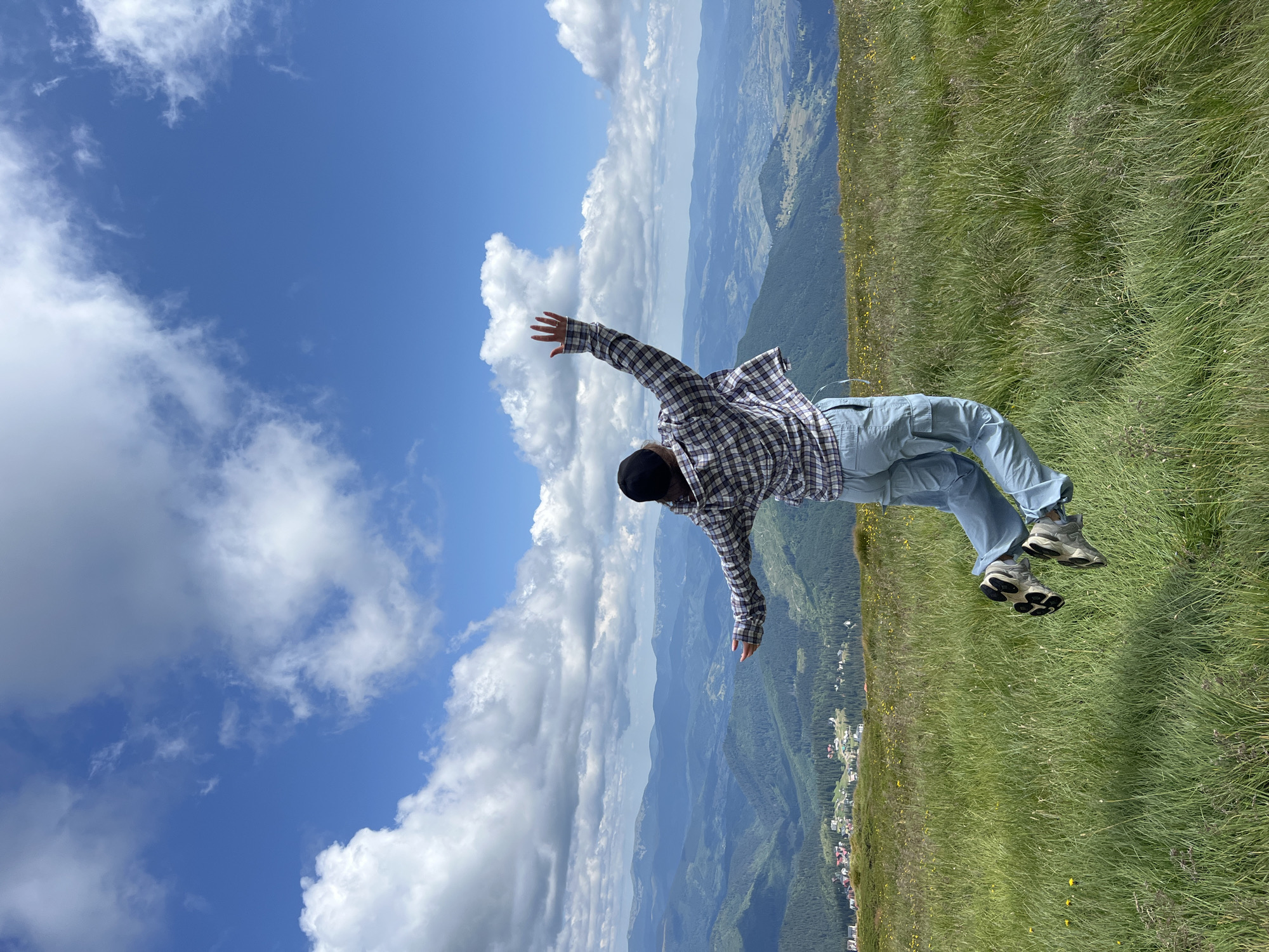 In the foreground, a person in a plaid shirt and light blue cargo pants is captured mid-jump in a grassy field. The person is wearing a dark baseball cap and athletic shoes. Their arms are outstretched, and their legs are bent. The background reveals a vast mountainous landscape with rolling green hills covered in trees. In the distance, a small village with red-roofed buildings is visible nestled among the hills. The sky above is a vibrant blue, dotted with fluffy white clouds. This scene is located in Chorna Tysa, Ukraine.