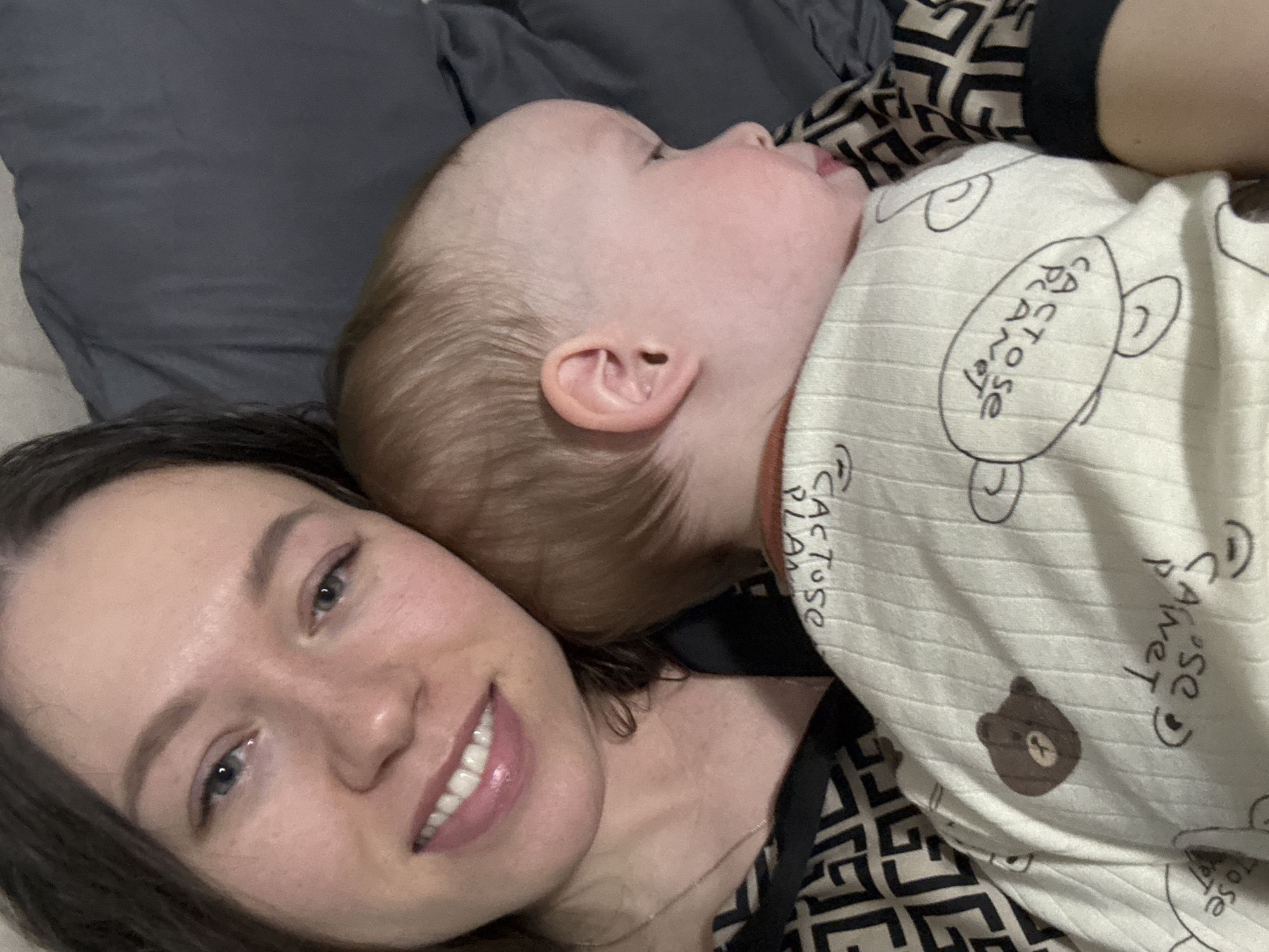 A woman is lying down with a baby sleeping on her chest. The woman has fair skin, dark hair, and is smiling. The baby is wearing a white patterned sleeper. They are lying on a bed with gray sheets and a patterned pillow. The location is potentially in the city of Ферма, Russia, based on the user prompt.