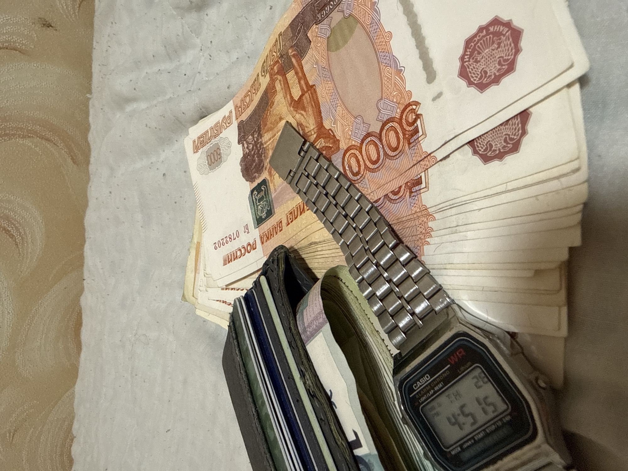The scene depicts a close-up of objects likely located in an indoor setting, possibly in an informal location rather than a bank. The primary objects in the foreground are a stack of Russian 5000 ruble banknotes, a men's digital Casio watch, and a leather wallet. The watch is placed on top of the stack of money, and the wallet is partially visible with cards inside. The environment is suggested to be a textured surface, likely a fabric material like a bed sheet or tablecloth. The time on the watch reads 4:51 and includes day information. The precise location cannot be determined beyond the presence of Russian currency, making a location in Лабжанскі сельскі Савет, Belarus, or any specific city within it, impossible to definitively confirm.