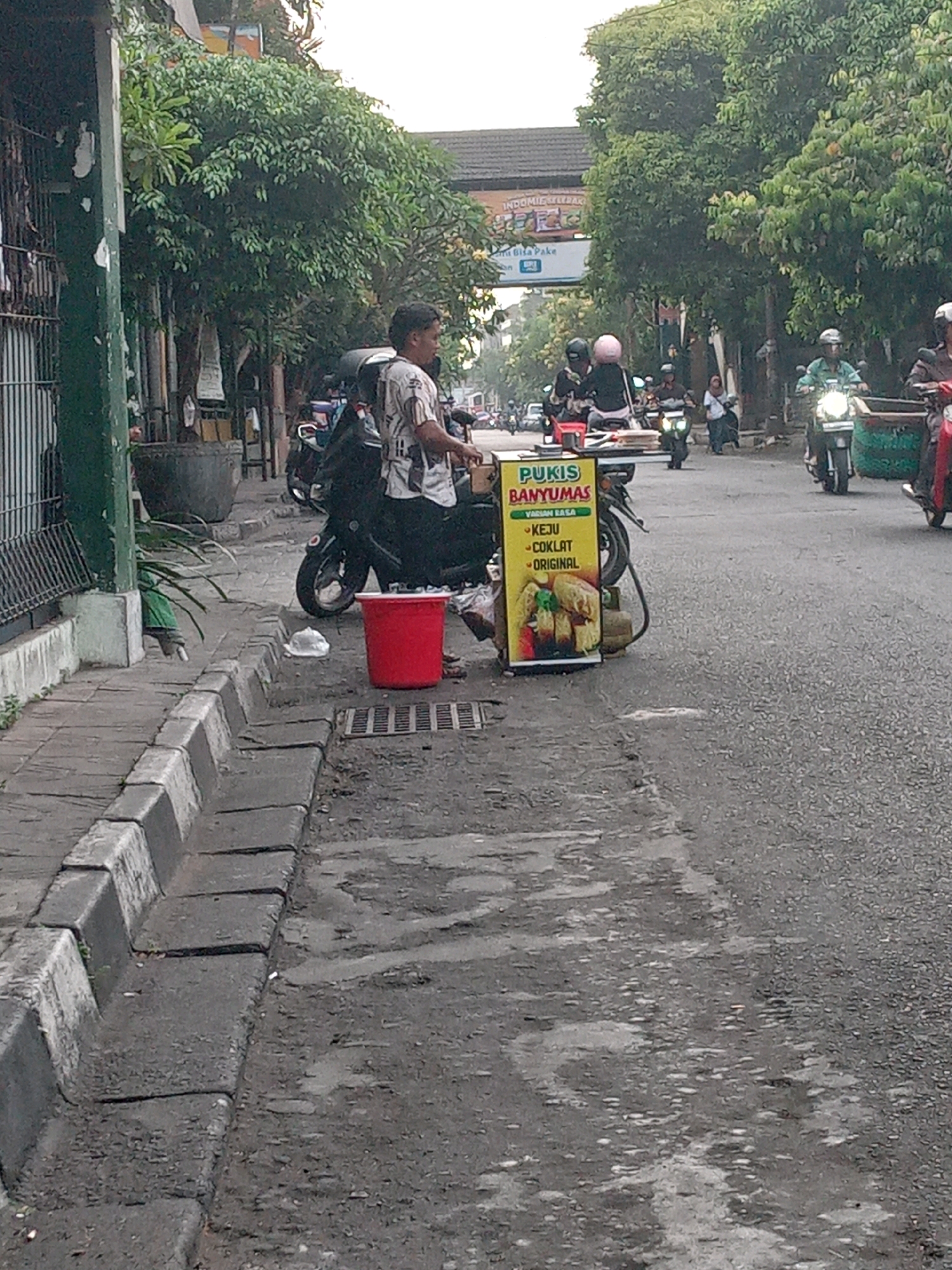 The image depicts a street scene in Yogyakarta, Indonesia, focusing on a vendor selling "Pukis Banyumas." A man, likely the vendor, stands behind a cart displaying various flavors of the snack, including "Keju," "Coklat," and "Original." The cart is a bright yellow with red lettering and images of the food. A red bucket is placed near the cart on the sidewalk. Several motorcycles are parked or being ridden on the street, indicating a typical urban environment. The background shows trees and buildings, consistent with a street in Yogyakarta. The overall scene appears to be an outdoor market or street-side commerce setting.
