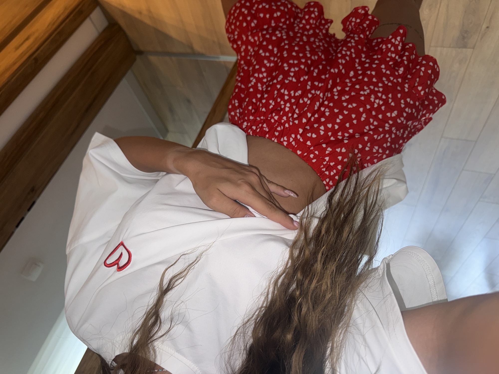 A high-angle shot shows a woman wearing a red and white heart-patterned top, a white t-shirt with a red heart logo, and white shoes. Her long, wavy brown hair is prominently displayed. The setting includes wooden furniture and what appears to be a bright interior with white walls. The location is Unknown, Unknown.