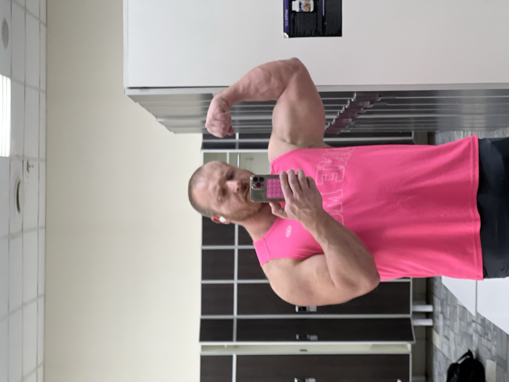 An adult male, with a shaved head and light facial hair, is captured from the waist up in what appears to be a gym locker room or changing area. He wears a bright pink sleeveless top and dark shorts. A white earbud is visible in his right ear. He holds a dark-colored smartphone with a textured case in his left hand, pointing it towards himself in a mirror selfie, while his right arm is bent, flexing his bicep. His facial expression is intense, with narrowed eyes. The background on the left features a row of dark-colored lockers with silver frames. On the right, lighter-colored lockers are visible, leading to a white wall adorned with a black poster advertising a supplement. The floor is tiled with a gray, patterned design, and a black bag rests on the floor to the left. The ceiling is a white grid with recessed lighting. The precise location within the city of Chaikovsky, Russia, is not identifiable from the image, though the setting is consistent with a fitness center.
