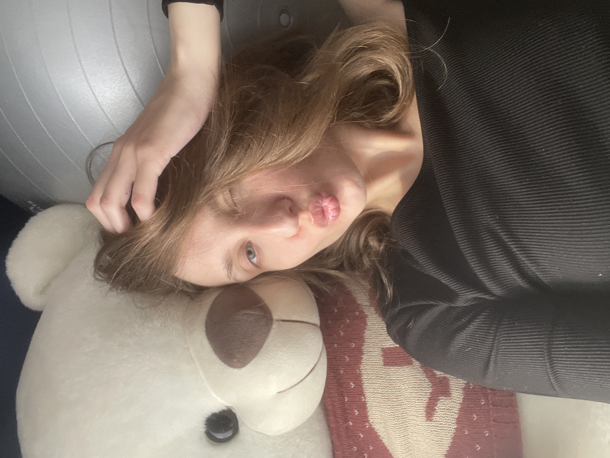 An indoor, close-up, diagonal portrait-style image captures a young person lying down, making a "kissy face" expression, with their head resting on a large white teddy bear. The subject has fair skin, light brown hair, and blue eyes, and is wearing a black ribbed long-sleeved top. Their left hand is visible, lightly touching their hair.
The large white plush teddy bear occupies the lower part of the frame, featuring dark button eyes and a brown nose. A patterned blanket or throw, with red/maroon and cream/beige tones, is visible beneath the person's right arm and adjacent to the teddy bear. In the upper left background, part of a light grey exercise or stability ball with distinctive ribbed lines is present. The lighting is soft, indicative of an indoor environment. The person's expression conveys a playful or affectionate mood. No text is visible in the image. The scene is set in Подшивалово, Russia.