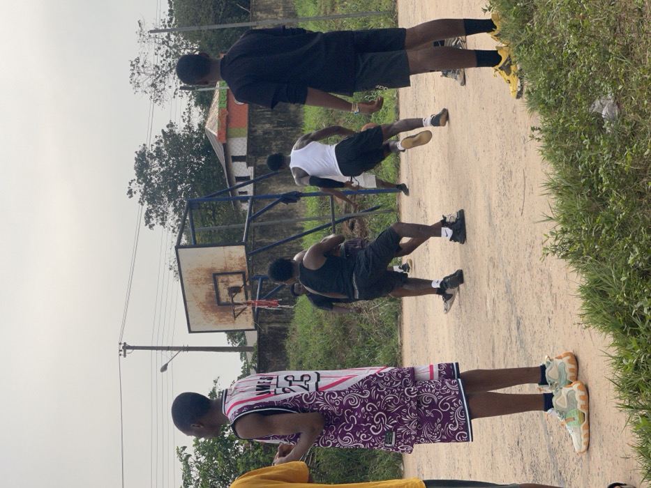 The image captures an outdoor basketball game in progress on a dirt court in Umunekwu, Nigeria. Six individuals are visible, primarily young men. In the foreground left, a person stands observing, wearing a white and purple patterned basketball jersey with "JAMES 23" on the back, and multi-colored sneakers. In the midground, four players are actively engaged in the game near a rusted basketball hoop with a white backboard and blue support frame. One player in a black tank top and black shorts is bent forward, while another in a white tank top and black shorts moves towards the right, away from the viewer. Other players are partially obscured. To the foreground right, another person stands watching, dressed in a black long-sleeved shirt, black shorts, and yellow and black sneakers. The ground consists of bare earth and sparse green vegetation. In the background, a utility pole with power lines is visible, along with a low concrete wall and distant buildings, one featuring a multi-colored roof. The sky is overcast.