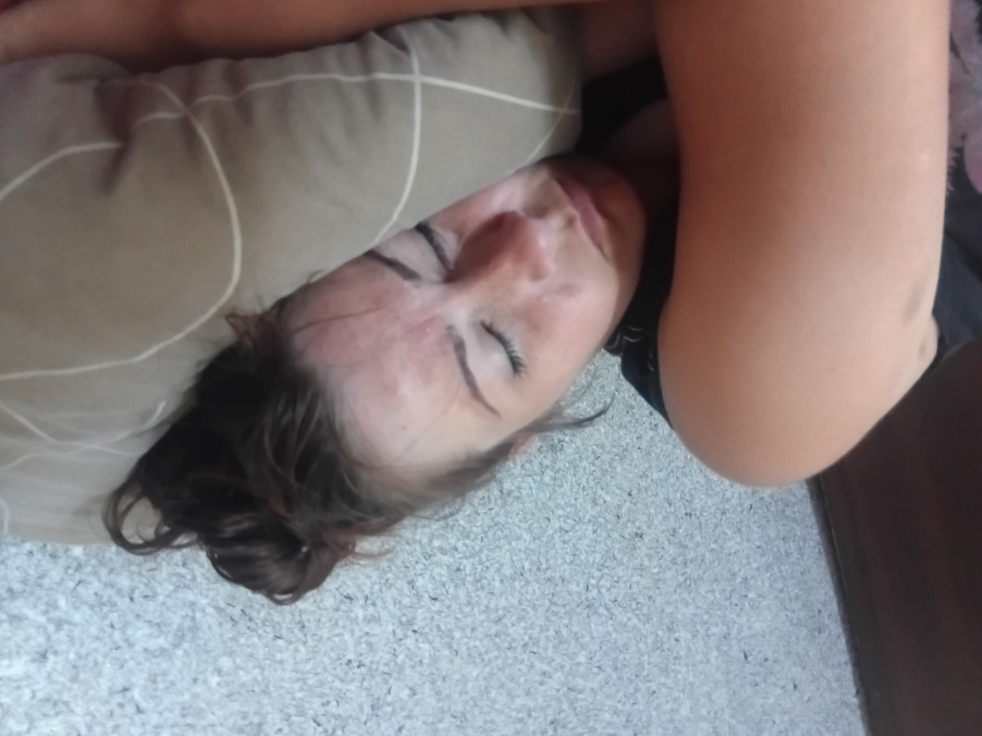 A person is lying down on a carpeted floor, with their head resting on a pillow. Their face is turned to the side and their eyes are closed. The person has dark hair and is wearing dark clothing. The scene is indoors, and the lighting suggests it could be daytime. The overall mood is peaceful and relaxed, suggesting the person might be sleeping or resting.