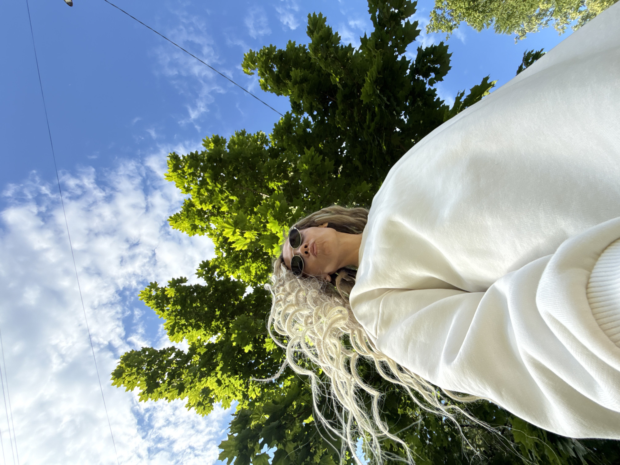 The image presents a low-angle, worm's-eye view of a person outdoors, captured against a vibrant blue sky with scattered white clouds. The subject, located in the upper right, is wearing a light-colored (likely white or off-white) sweatshirt and round, dark sunglasses. Their long, light-blonde or heavily highlighted curly hair cascades downwards, appearing voluminous and textured. The person is making a slight pouting expression.
Lush green tree foliage, possibly from maple trees given the shape of the leaves, fills the left and lower parts of the frame, bathed in sunlight. Thin, dark overhead wires stretch across the sky, suggesting an urban or suburban outdoor setting. The perspective implies the photo was taken by someone looking up, possibly a selfie. The time of day is clearly daytime, with bright conditions. No visible text is present. The scene is set in Rusanovo, Russia.