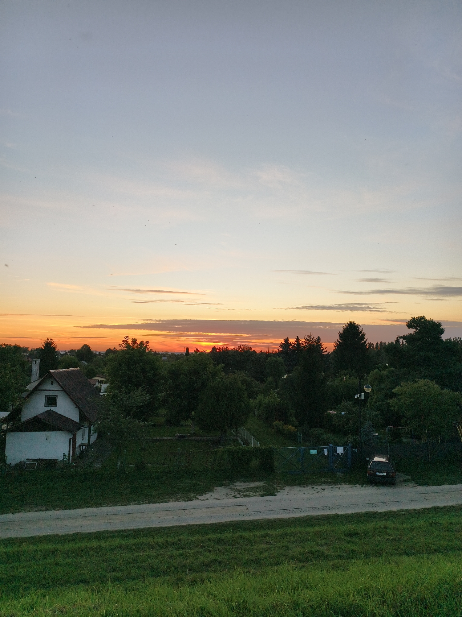 The image depicts a rural or suburban landscape at sunset. The sky is a gradient of pale blue to orange, with thin clouds stretching horizontally across the horizon. In the mid-ground, there is a dense array of trees, with a white house and its chimney visible on the left side, partially obscured by foliage. A dirt road runs horizontally across the lower mid-ground, with a dark-colored car parked near a fenced area on the right. The foreground consists of a grassy embankment. The scene appears to be located in a quiet, undeveloped area, possibly on the outskirts of a town or city. Based on the context provided that this image is from Tczew, Poland, it is likely a view of the outskirts of that city, potentially in a rural or semi-rural area with scattered residential properties. No people are visible in the image.