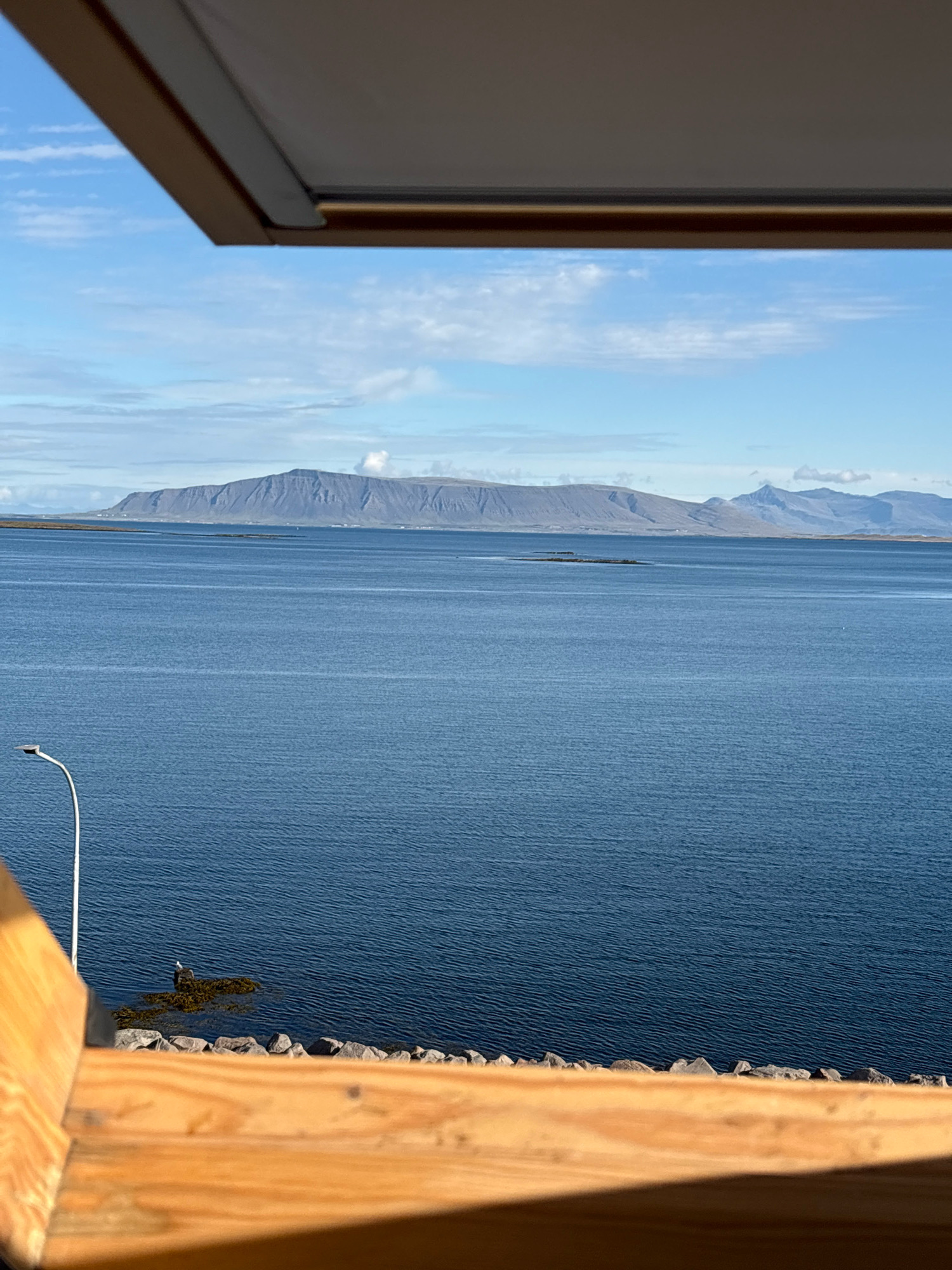 The image shows a view of the ocean from a wooden structure, likely a balcony or deck. In the distance, a long, mountainous island is visible across the water, under a partly cloudy blue sky. In the foreground, a portion of the wooden structure occupies the bottom and top edges of the frame, casting a shadow across the lower part. A single white lamppost stands on the left side of the image, near a rocky shoreline. A small bird is perched on a cluster of rocks in the water, just to the left of the lamppost. The overall scene suggests a coastal location in Reykjavik, Iceland, with a calm body of water stretching towards the distant mountains.