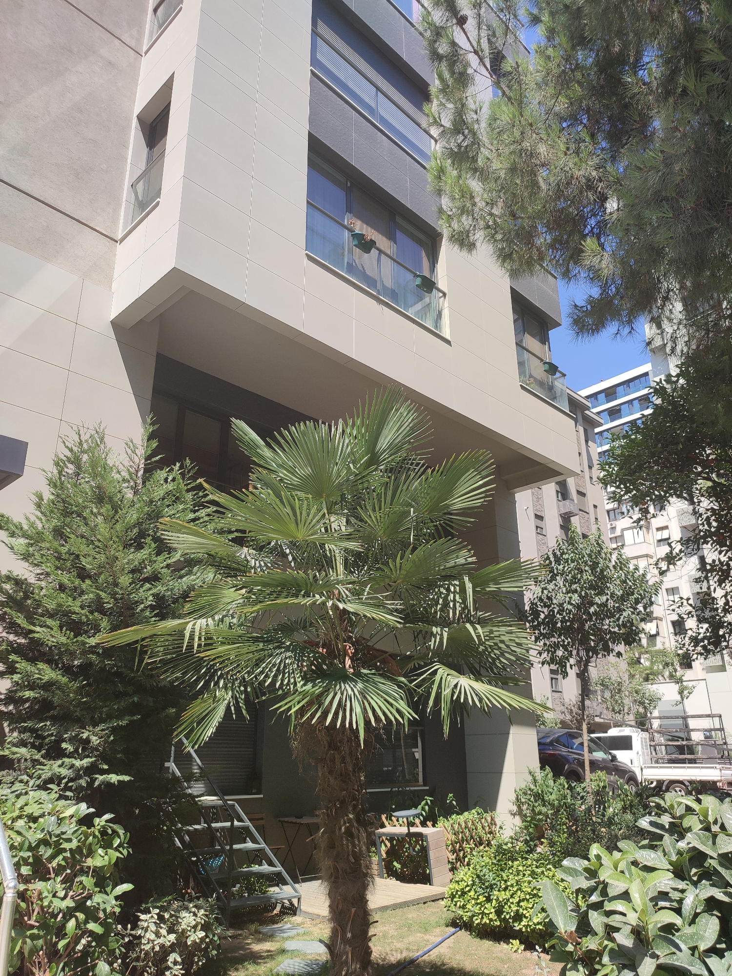 The image shows a modern apartment building with a grey facade and large windows, located in Kadıköy, Turkey. In the foreground, there are various plants, including a palm tree, a conical evergreen tree, and dense green bushes. To the right of the building, another apartment building is visible, featuring a blue-tinted glass facade. A dark-colored car and a white truck are parked in the background, near the entrance of the building. There are no people visible in the scene. The environment appears to be an urban setting with a mix of architecture and greenery.
