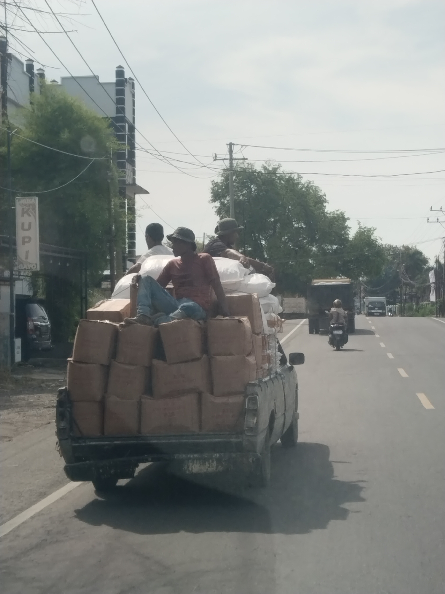 The image shows a gray pickup truck heavily laden with brown cardboard boxes on a road in Bireuen, Indonesia. Three men are sitting atop the load, two in hats, and one with blue jeans. The truck is traveling on a two-lane road with a broken yellow center line. A motorcyclist is riding behind the truck, and further down the road, a truck and a van are visible. On the left side of the road, there is a building with a sign that reads "KUPI GAYO." Trees line both sides of the road, and power lines are visible overhead. The sky is overcast.