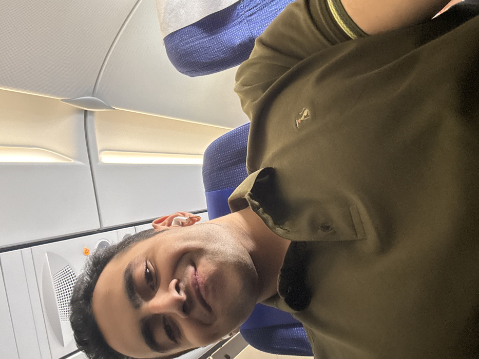 A man is sitting in an airplane seat. The interior of the airplane is visible, including the overhead compartments, lighting, and the blue seat in front of him. The man is wearing a green polo shirt with a small logo on the chest. He is looking at the camera and smiling slightly. The image is rotated, with the man's head near the bottom of the frame.