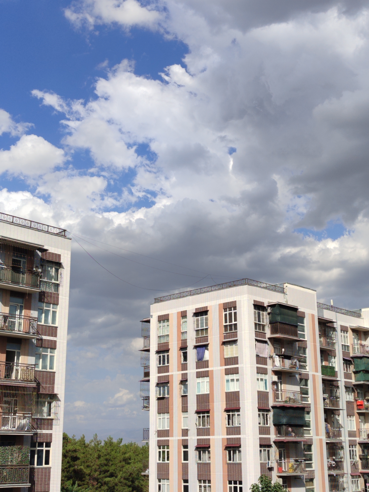 The image captures an outdoor scene featuring two apartment buildings against a backdrop of a partly cloudy sky. The buildings are tall, multi-story structures with balconies on each level. The building on the left appears to be closer to the viewer, while the building on the right is situated further back.
The apartment building on the left is characterized by its light-colored exterior, possibly white or light grey, adorned with decorative tilework in shades of brown and possibly maroon around the windows and balconies. The balconies themselves are enclosed with metal railings and some are fitted with awnings or screens. There are visible window panes, some of which appear to be open, and laundry hanging on clotheslines on some of the balconies.
The apartment building on the right shares a similar architectural style, with light-colored facades and balconies. It also displays decorative tile accents in complementary colors. Visible on its balconies are awnings and railings, and some windows are apparent.
Between the two buildings, a line, possibly a clothesline or communication wire, is stretched across the sky, connecting the structures. Below the buildings, a dense line of green trees is visible, suggesting the presence of a park, forest, or a residential area with significant greenery.
The sky is a mix of blue and grey clouds. Patches of bright blue sky are interspersed with large, fluffy white clouds and darker, more ominous grey clouds, suggesting dynamic weather conditions.
No people are clearly discernible in the image, nor are there any explicit actions or interactions being captured. The scene is a static view of urban architecture and the natural sky. The context provided about Minglola mahalla, Uzbekistan, situates this image within a specific geographical and cultural setting, implying the architectural style might be typical of residential buildings in that region.